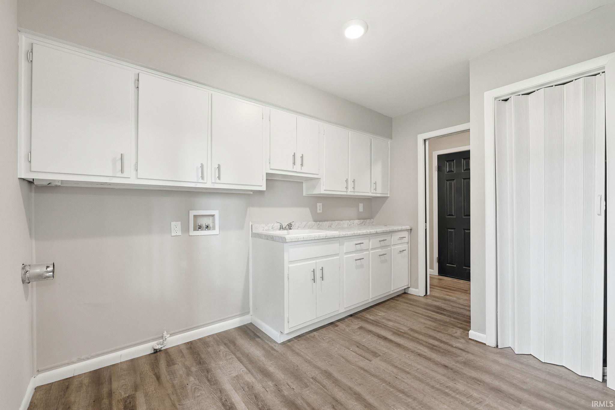 Spacious utility room featuring extensive white cabinetry, a built-in sink, wood-finish flooring, and a folding accordion door