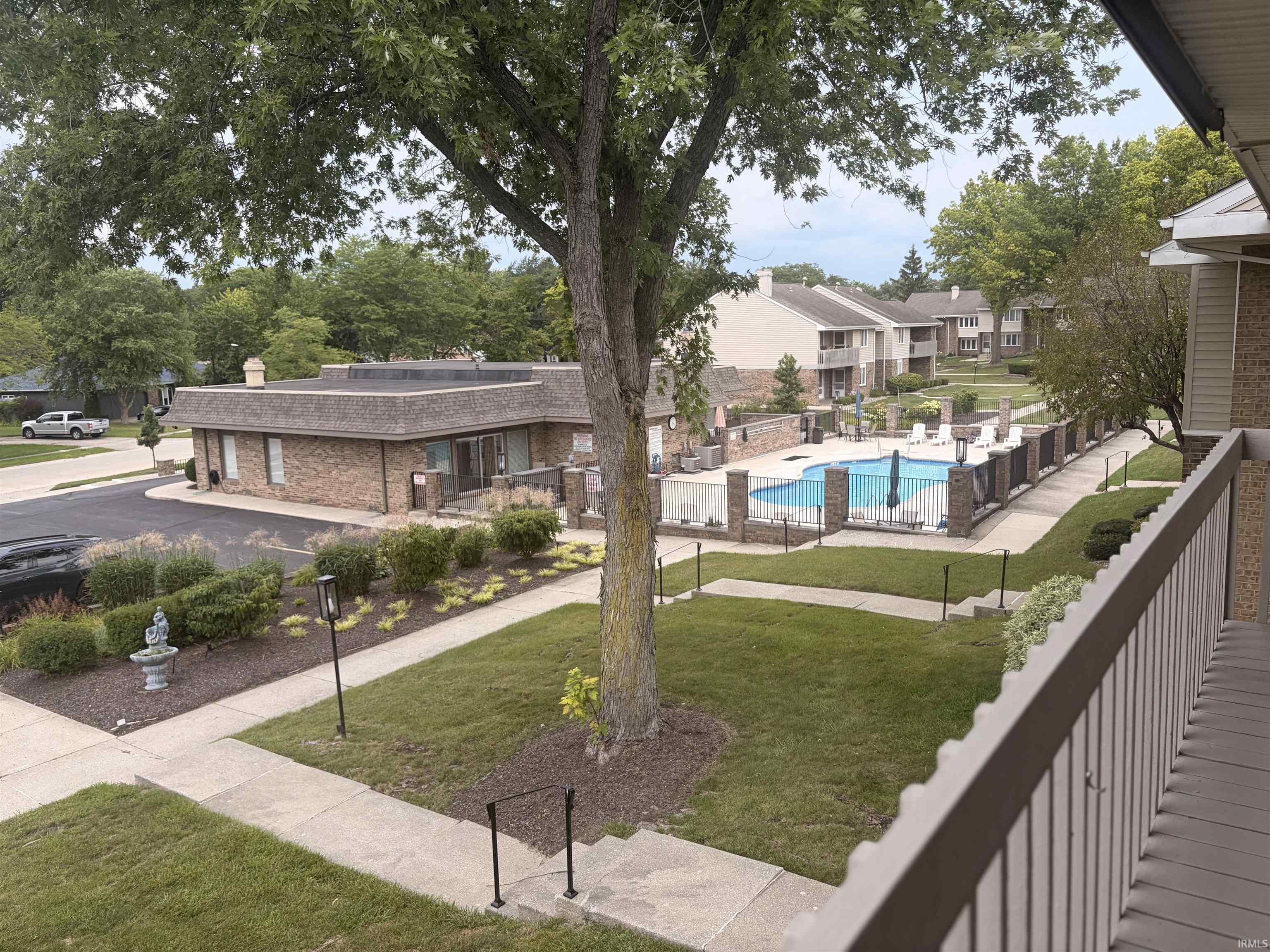Outdoor swimming pool with surrounding fence and paved patio, extensive landscaping with manicured lawns and mature trees, and a brick-faced building with a shingled roof