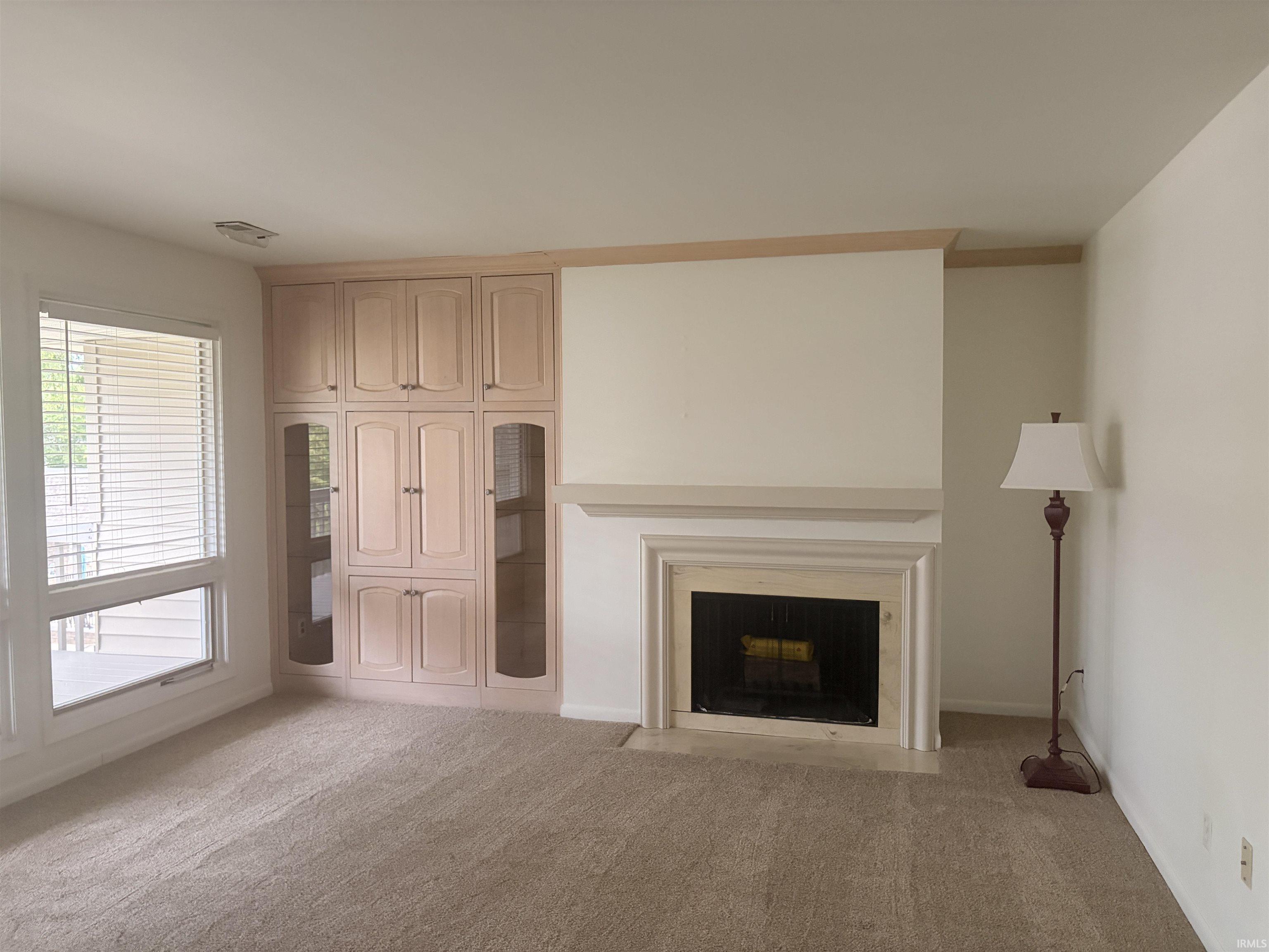 Built-in cabinetry with glass-front display, large window with blinds, fireplace with a mantel, and neutral carpeting