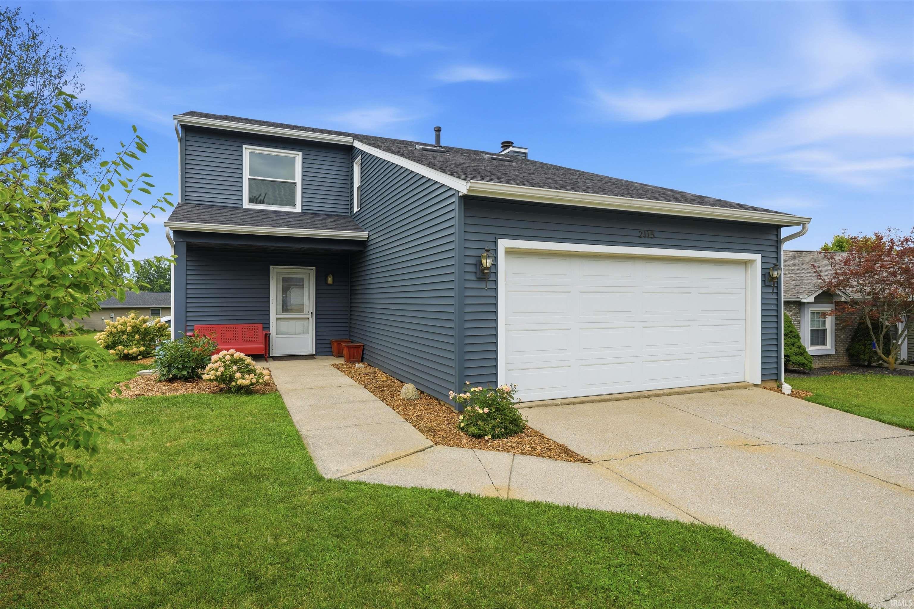 Two-story home featuring blue horizontal siding, a two-car attached garage with white paneled door, concrete driveway, and a manicured front lawn with mature landscaping and flowering shrubs