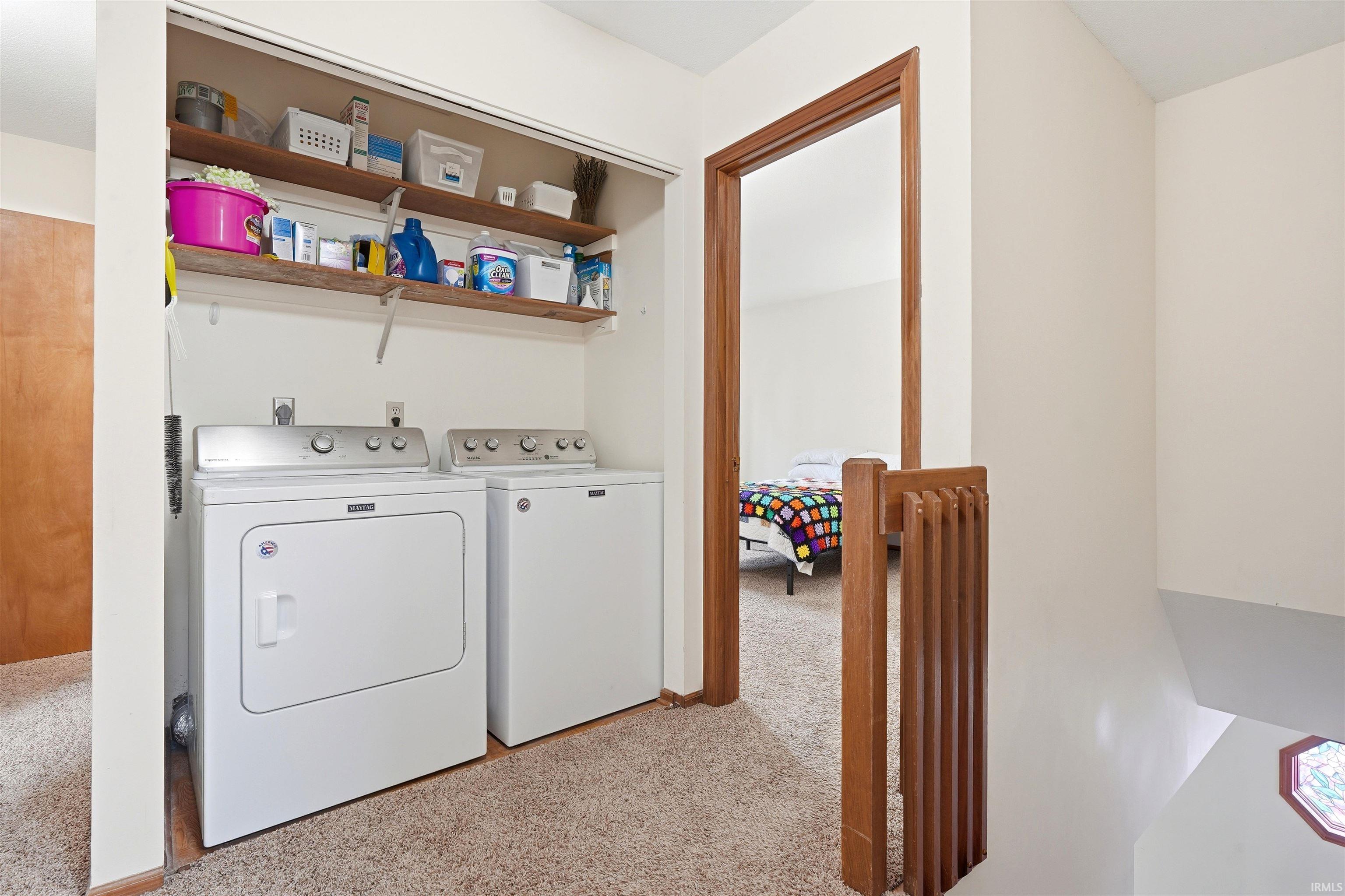 Dedicated laundry room featuring side-by-side washer and dryer appliances, open shelving with storage organization, and carpeted flooring