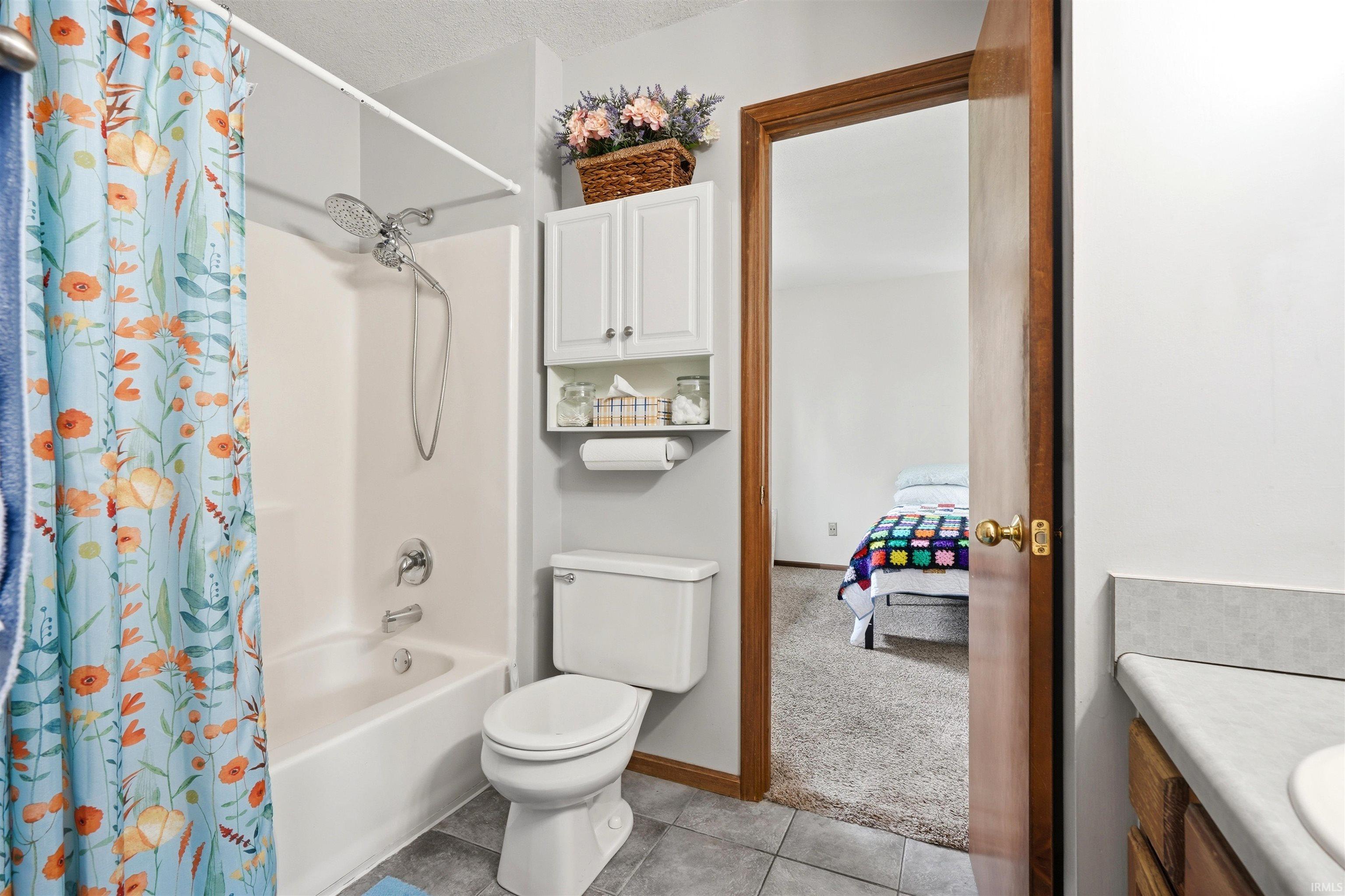 Full bathroom featuring a white porcelain toilet, bathtub with shower surround and colorful floral curtain rod, ceramic tile flooring, white cabinetry with open shelving, and wood-finish framed doorway providing access to the adjoining bedroom