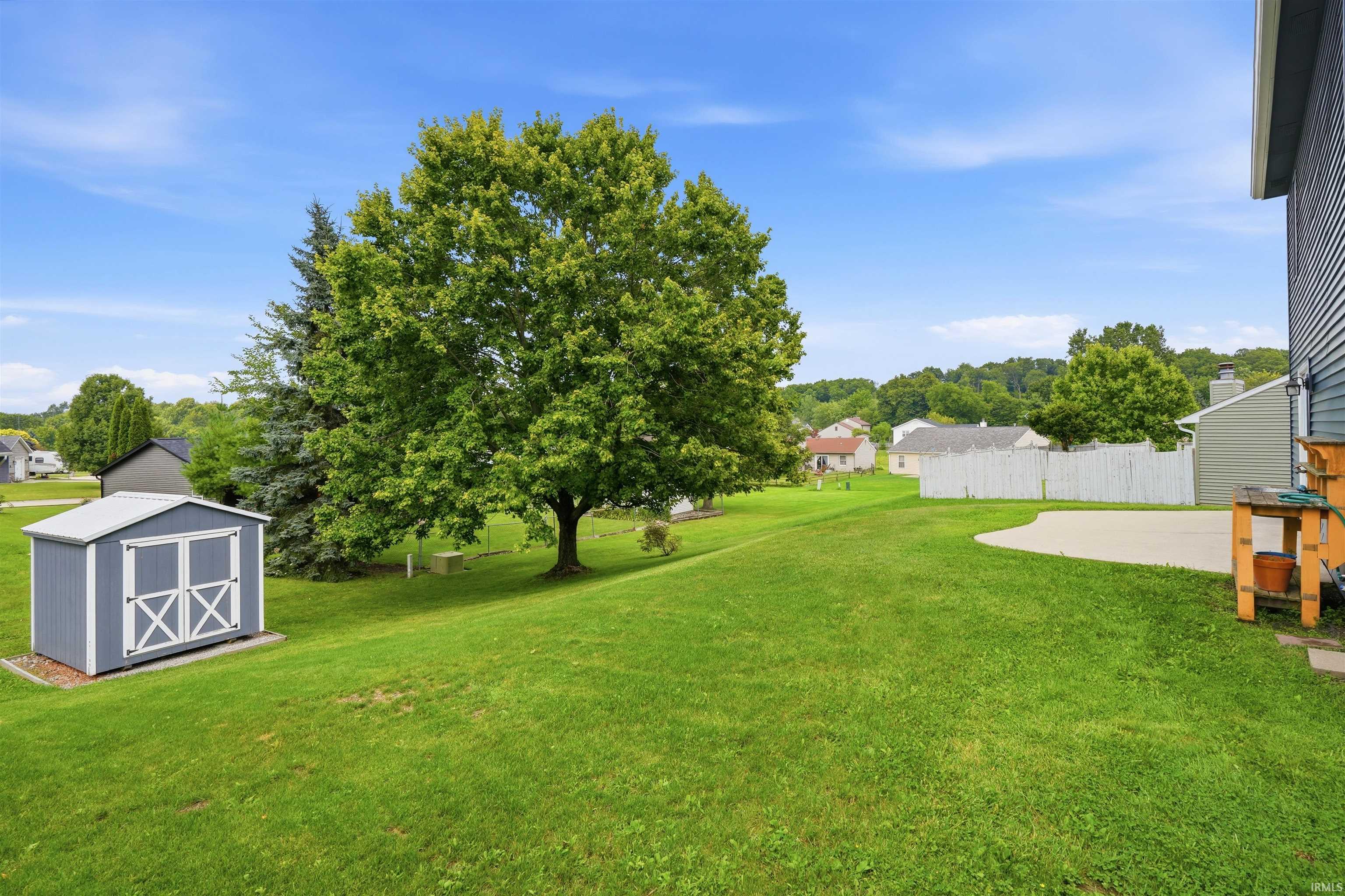 Expansive level grounds featuring mature shade trees and lush green lawn, complemented by a detached storage shed with X-pattern doors and a single-story residence situated on the right