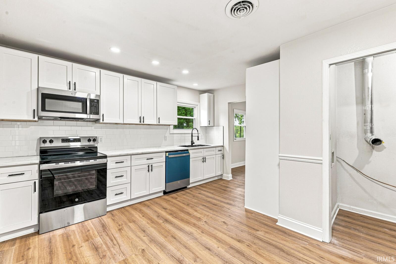 Kitchen featuring white shaker cabinetry, stainless steel appliances, white subway tile backsplash, light-toned countertops, and wood-finish flooring