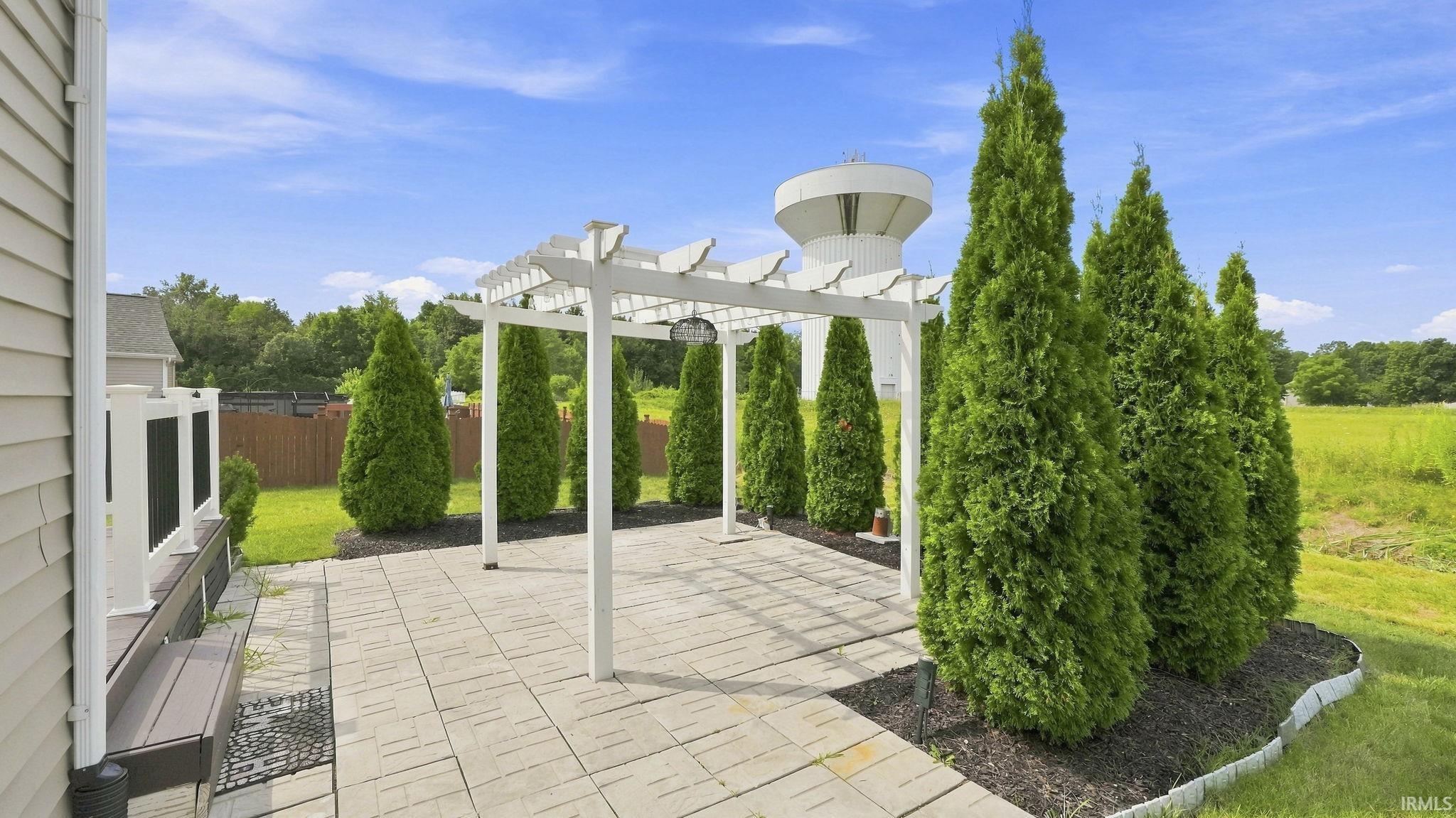 Expansive paved patio with a white pergola, surrounded by mature evergreen trees