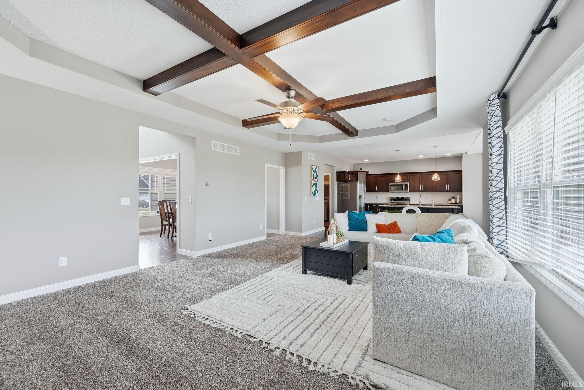 Open-concept living area with coffered ceiling featuring dark wood beams, ceiling fan with brushed metal finish, and abundant natural light from expansive windows with plantation shutters
