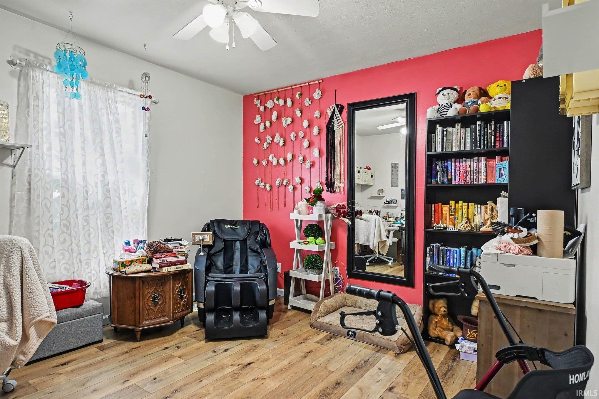 Wood-finish flooring, ceiling fan with integrated lighting, window with sheer curtains, a full-length mirror, and a built-in bookshelf