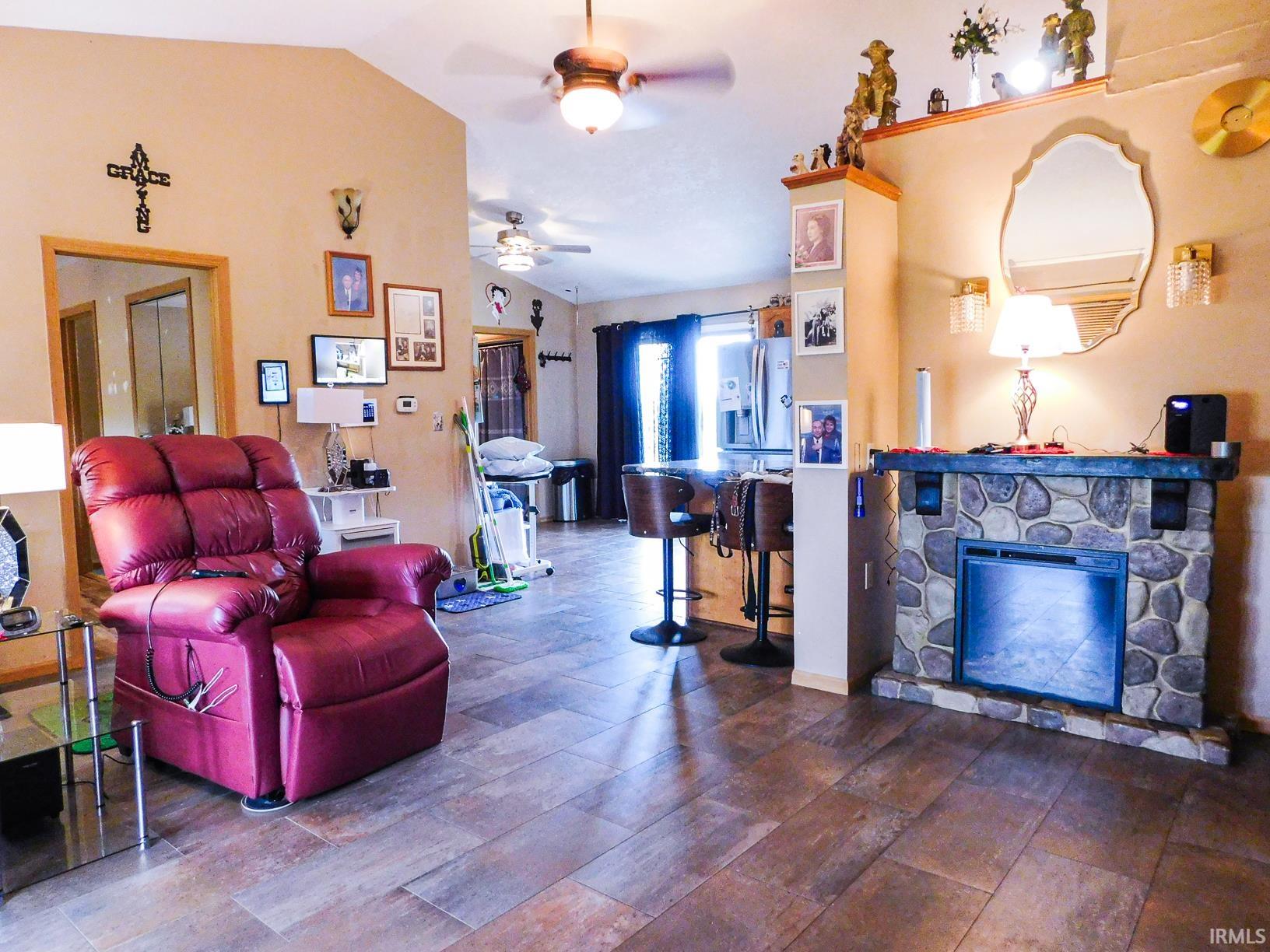 Vaulted ceiling, wood-finish flooring, stone-clad fireplace, ceiling fan, and recessed kitchen area with bar seating