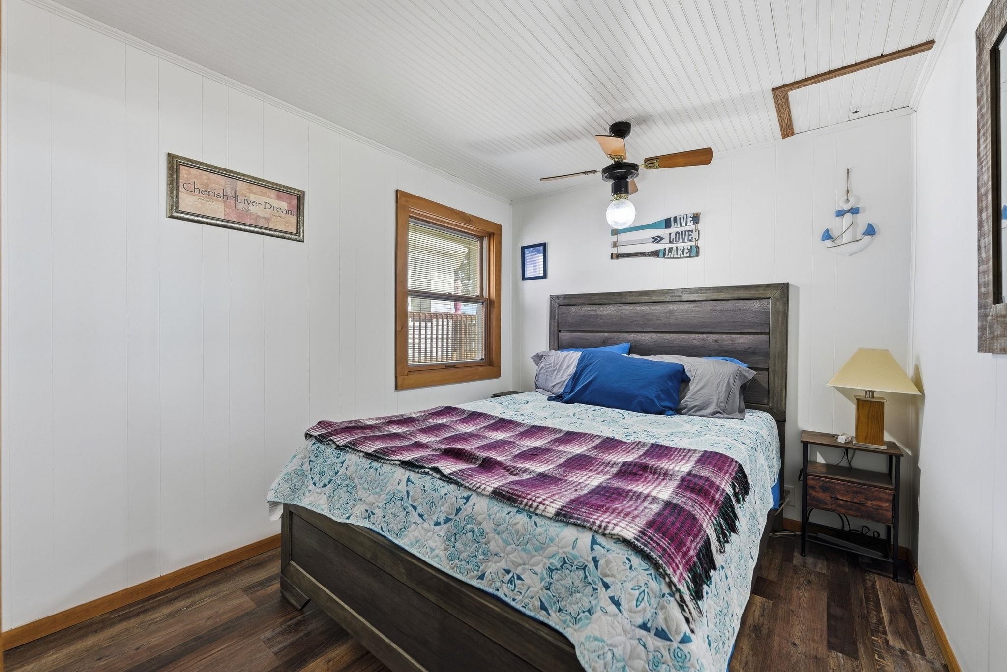 Comfortable bedroom featuring a wood-finish flooring, charcoal-upholstered bed frame, ceiling fan with wood blades, and bedside nightstand with drawer storage