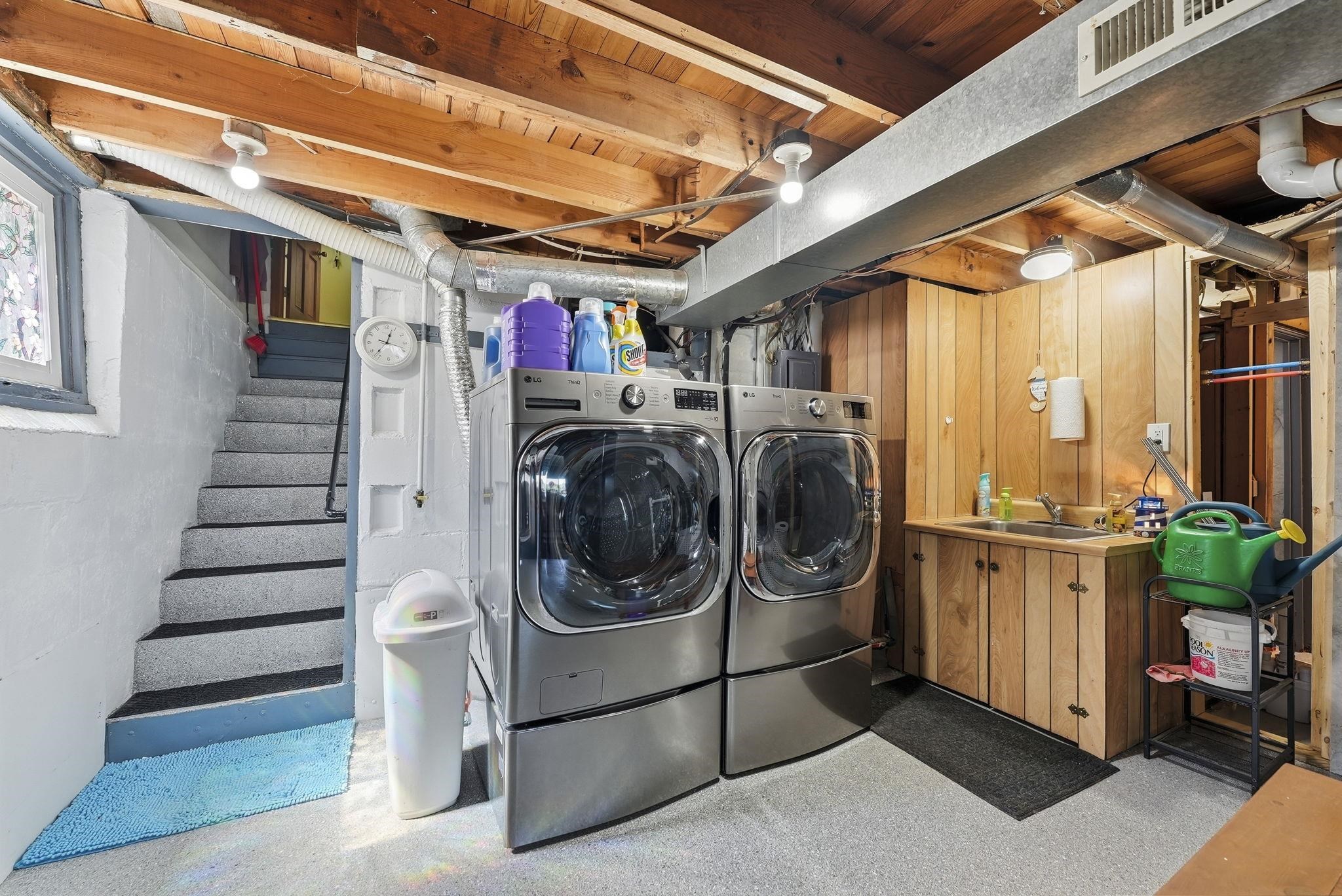 Finished Basement Laundry Room featuring high-capacity front-load washer and dryer with pedestal bases, wood-finish cabinetry with ample storage, epoxy-coated concrete flooring, exposed wood beam ceiling, and direct access to stairs