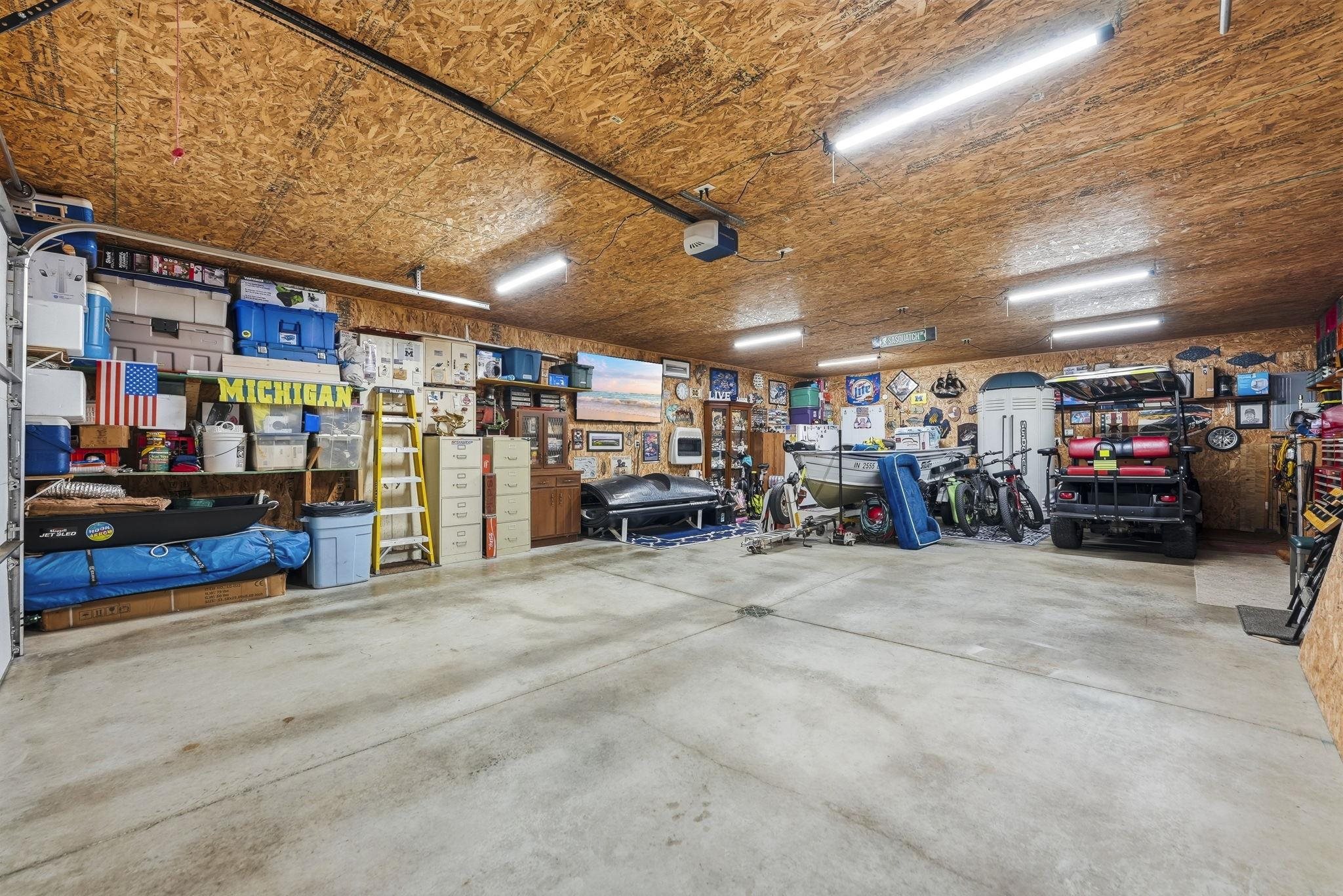 Spacious garage featuring a concrete floor, exposed wood ceiling, multiple fluorescent light fixtures, and extensive built-in shelving