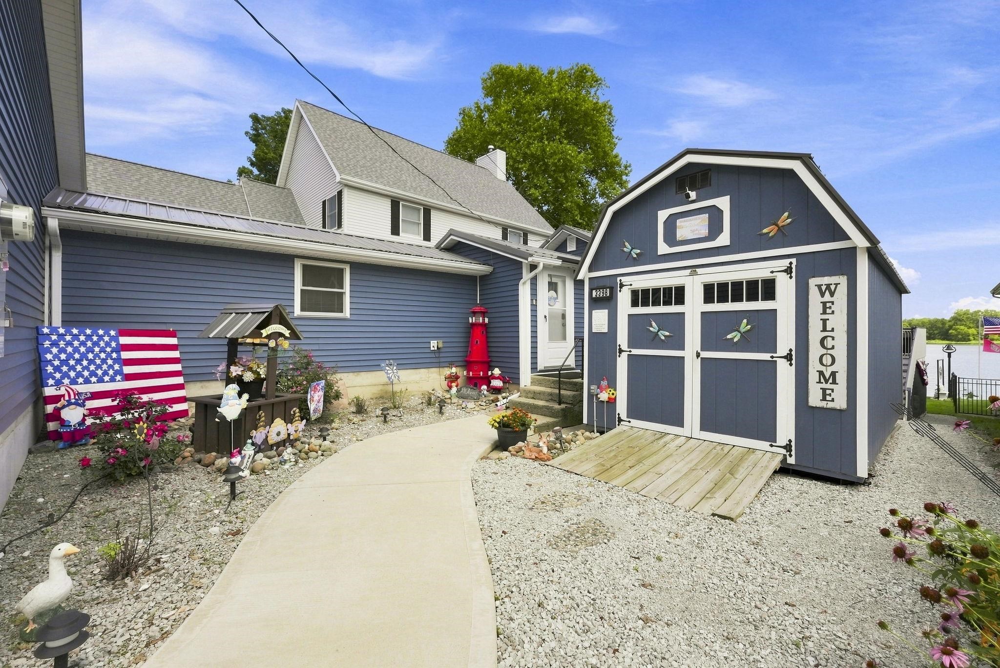 Blue siding, gambrel-roofed shed with double doors, concrete pathway, gravel ground cover, and landscaping with perennial plants