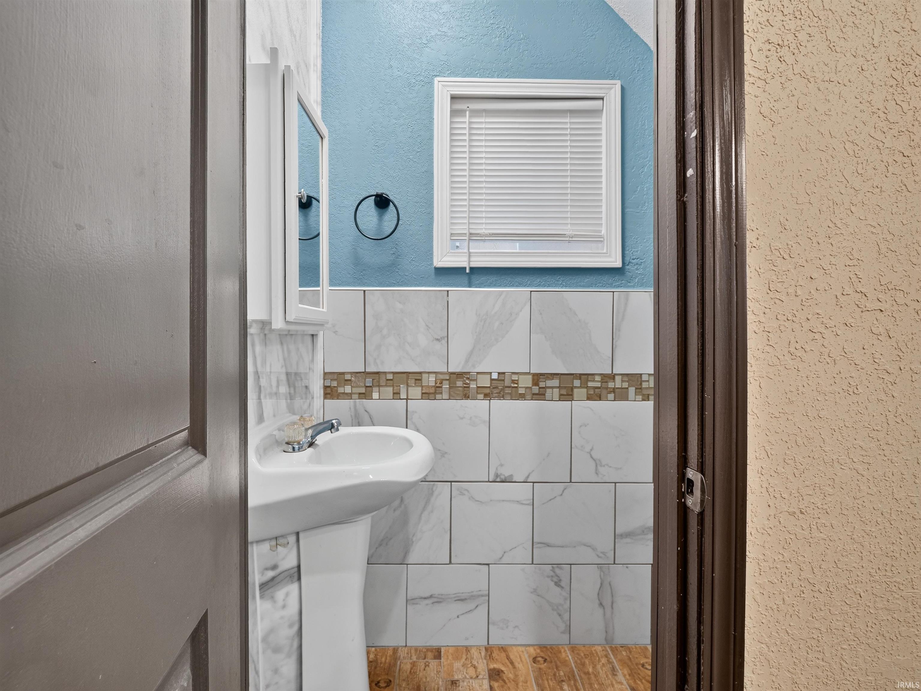 Pedestal sink, wall-mounted medicine cabinet with mirror, tiled wall with decorative mosaic band, wood-finish flooring, and window with blinds