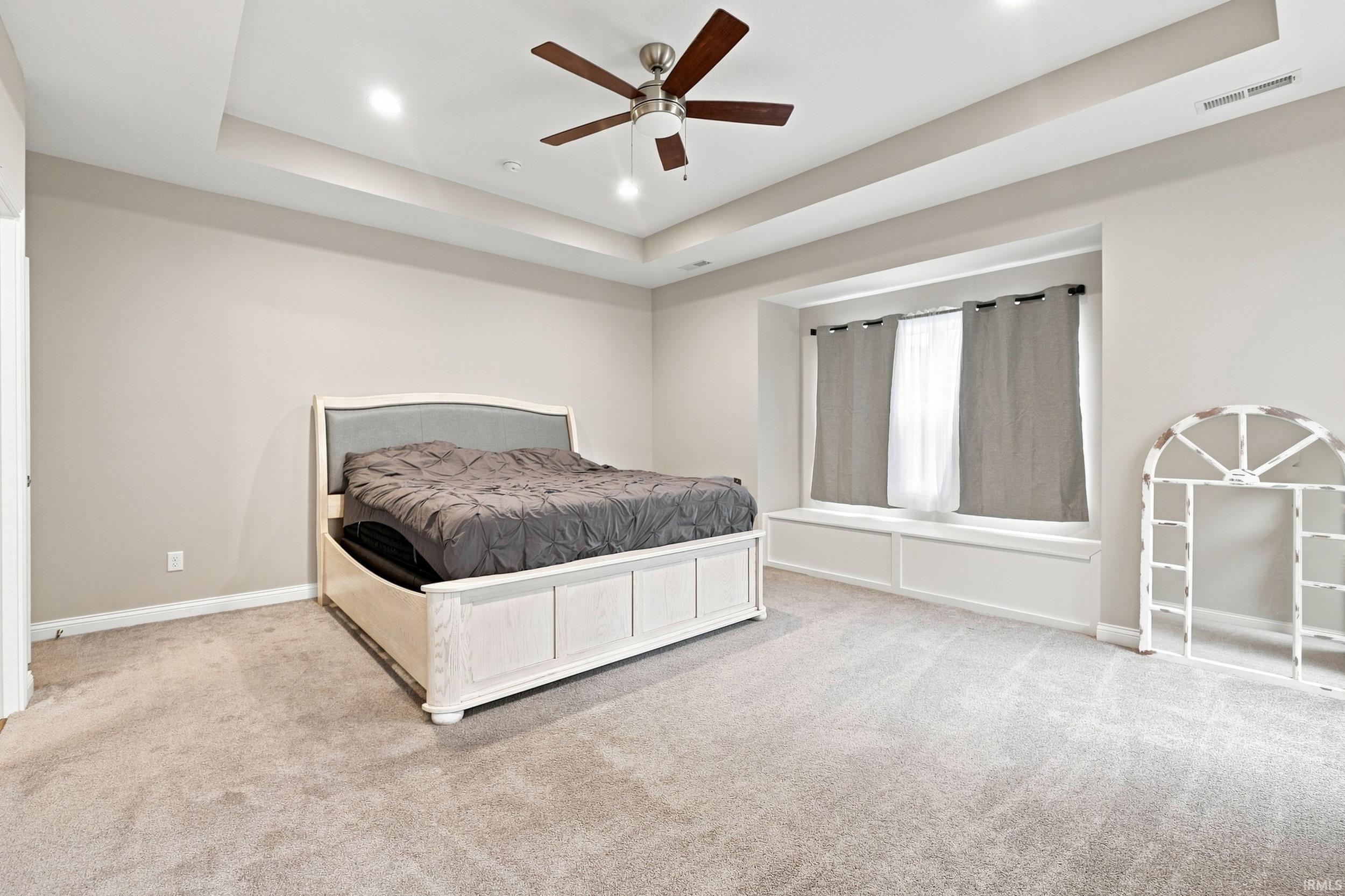 Spacious primary bedroom featuring a coffered ceiling with ceiling fan, expansive windows with neutral drapery, neutral carpet flooring, and abundant natural light from multiple glass doors