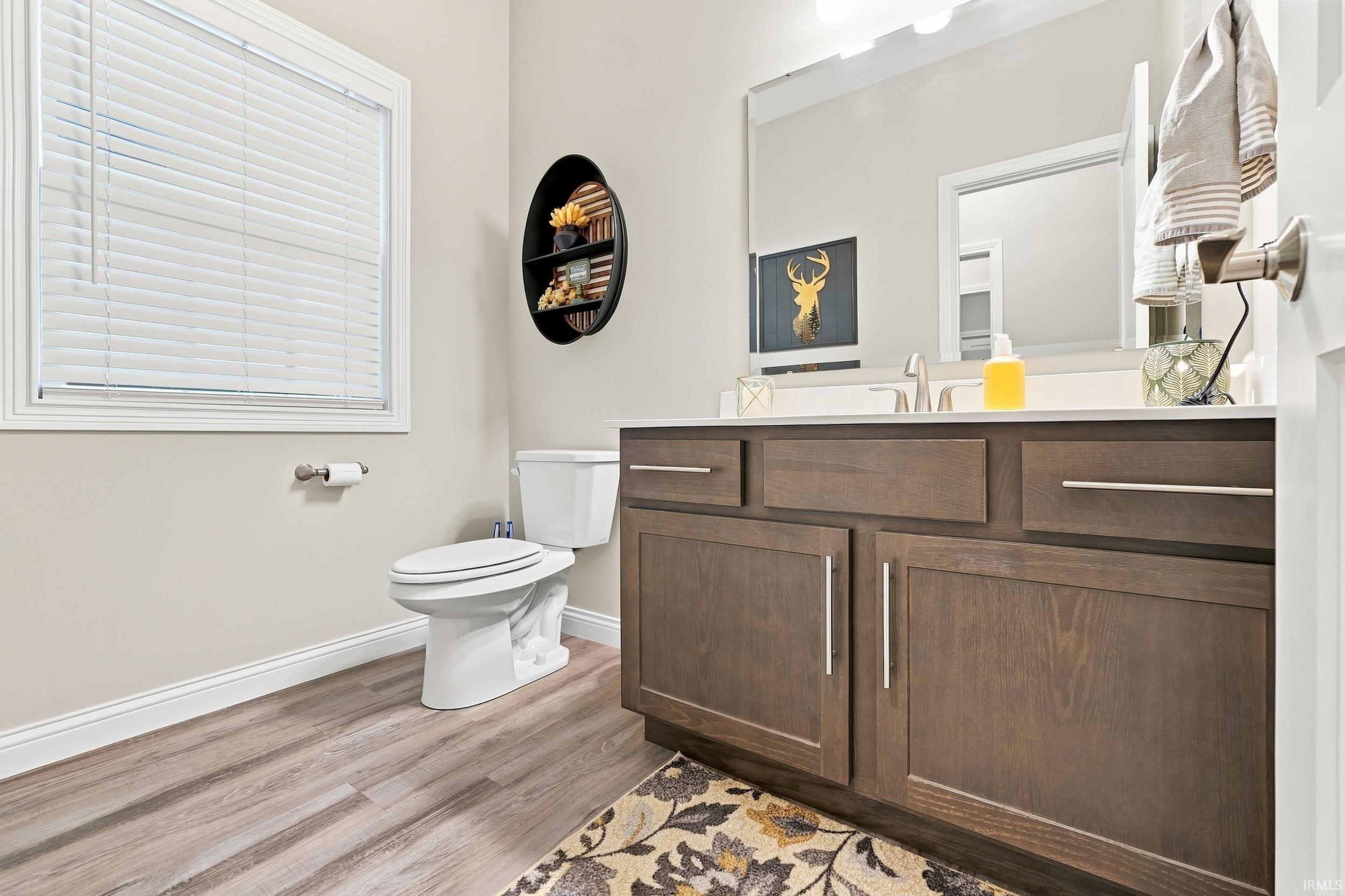 Wood-finish flooring, white window with blinds, white toilet, dark wood vanity with white countertop, and large wall-mounted mirror