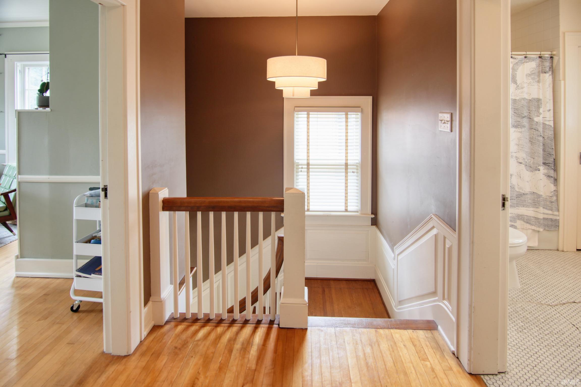 Hardwood flooring, painted white trim, white window blinds, tiered chandelier, and a white stair railing with wood-finish handrail
