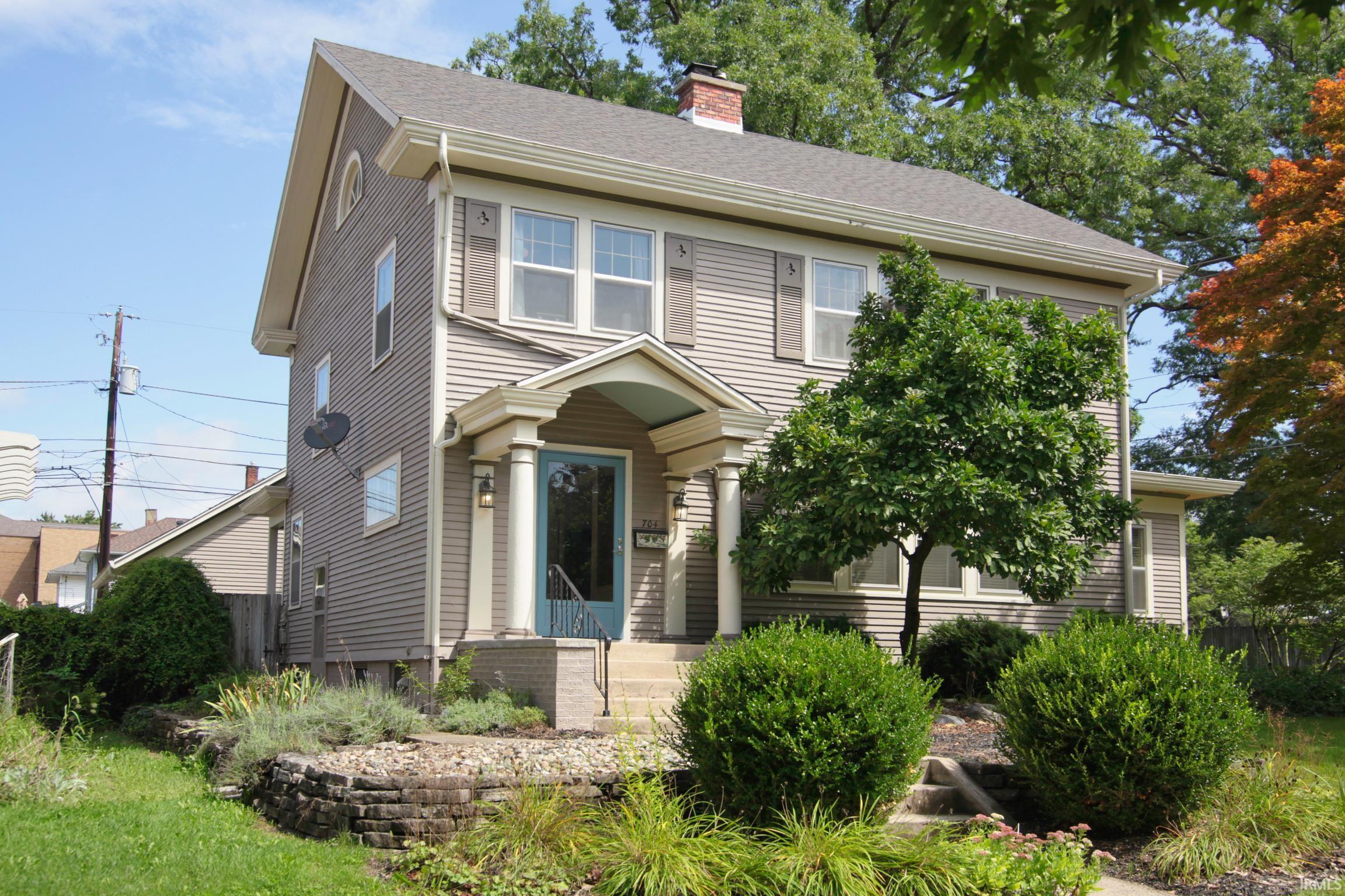Siding exterior with decorative shutters, covered porch with column supports, established landscaping, and a brick chimney