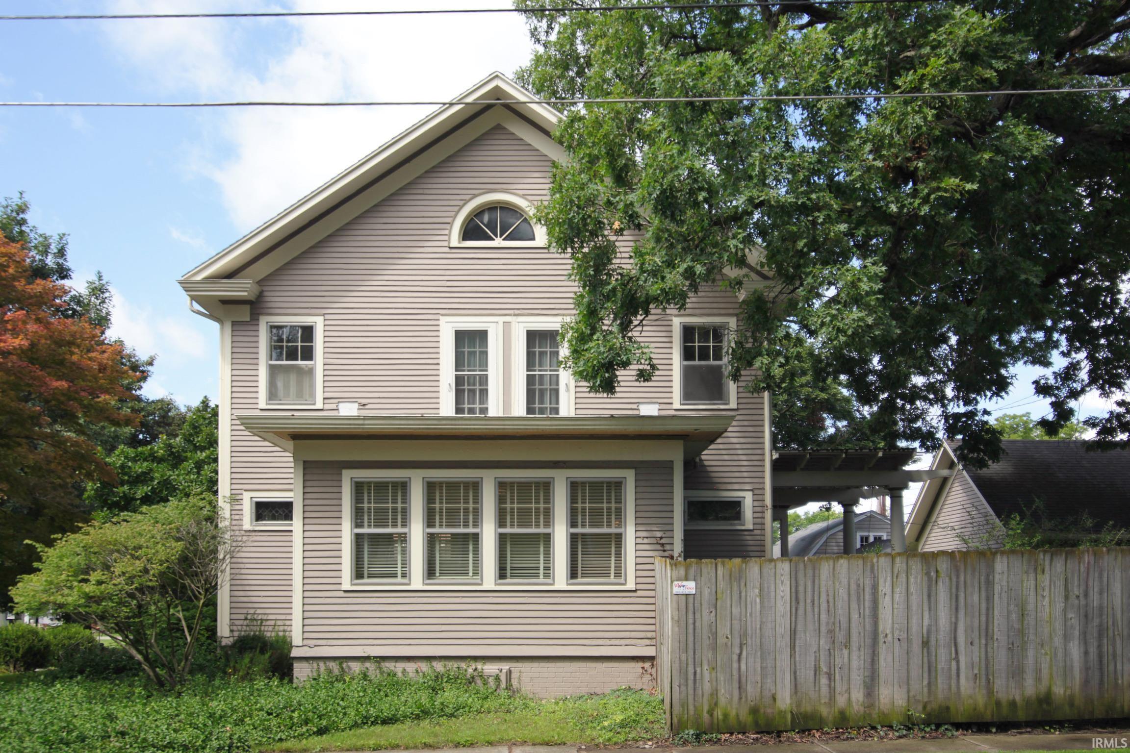 Gable roofline with an arched window, horizontal siding exterior, multiple pane windows, mature perimeter landscaping, and a wooden fence
