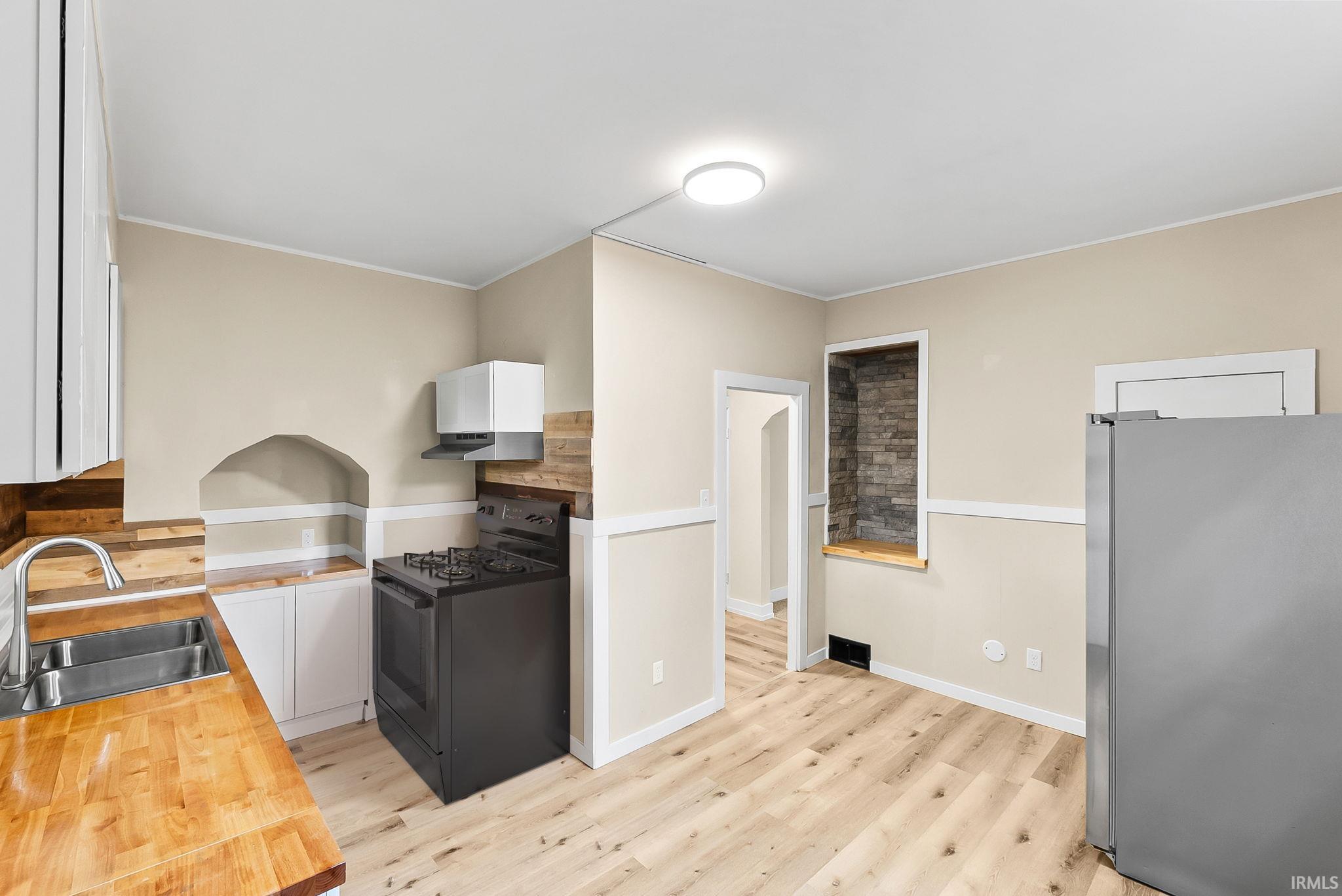 Light wood-finish flooring, butcher block countertops, stainless steel double basin sink, modern ceiling light fixture, and white cabinetry