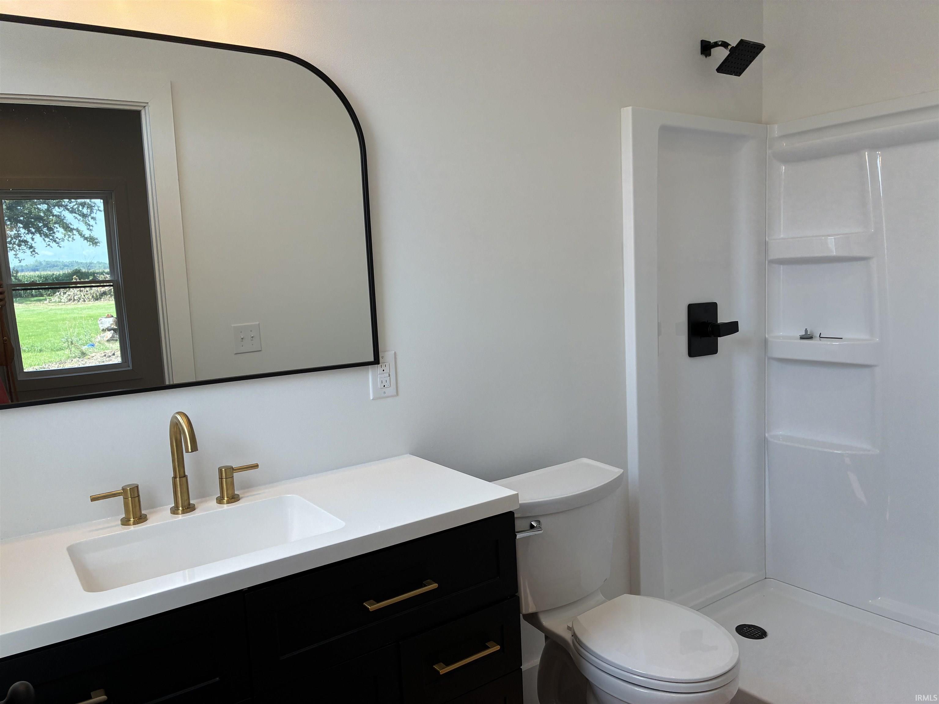 Vanity with integrated rectangular sink and gold-tone faucet, black framed arch mirror, black vanity cabinet with gold-tone pulls, white shower surround, and matte black shower fixtures