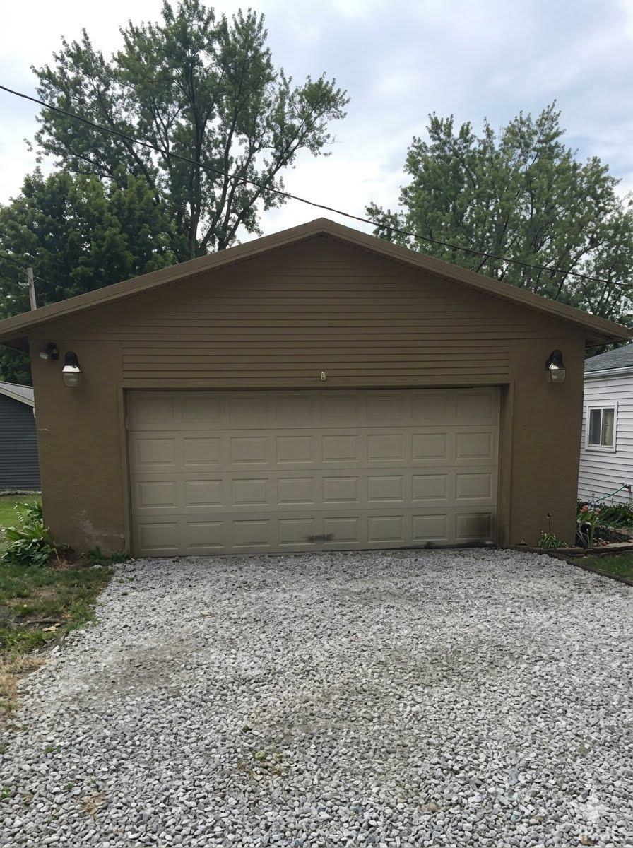 Detached garage with a paneled overhead door, gravel driveway, and exterior wall sconces