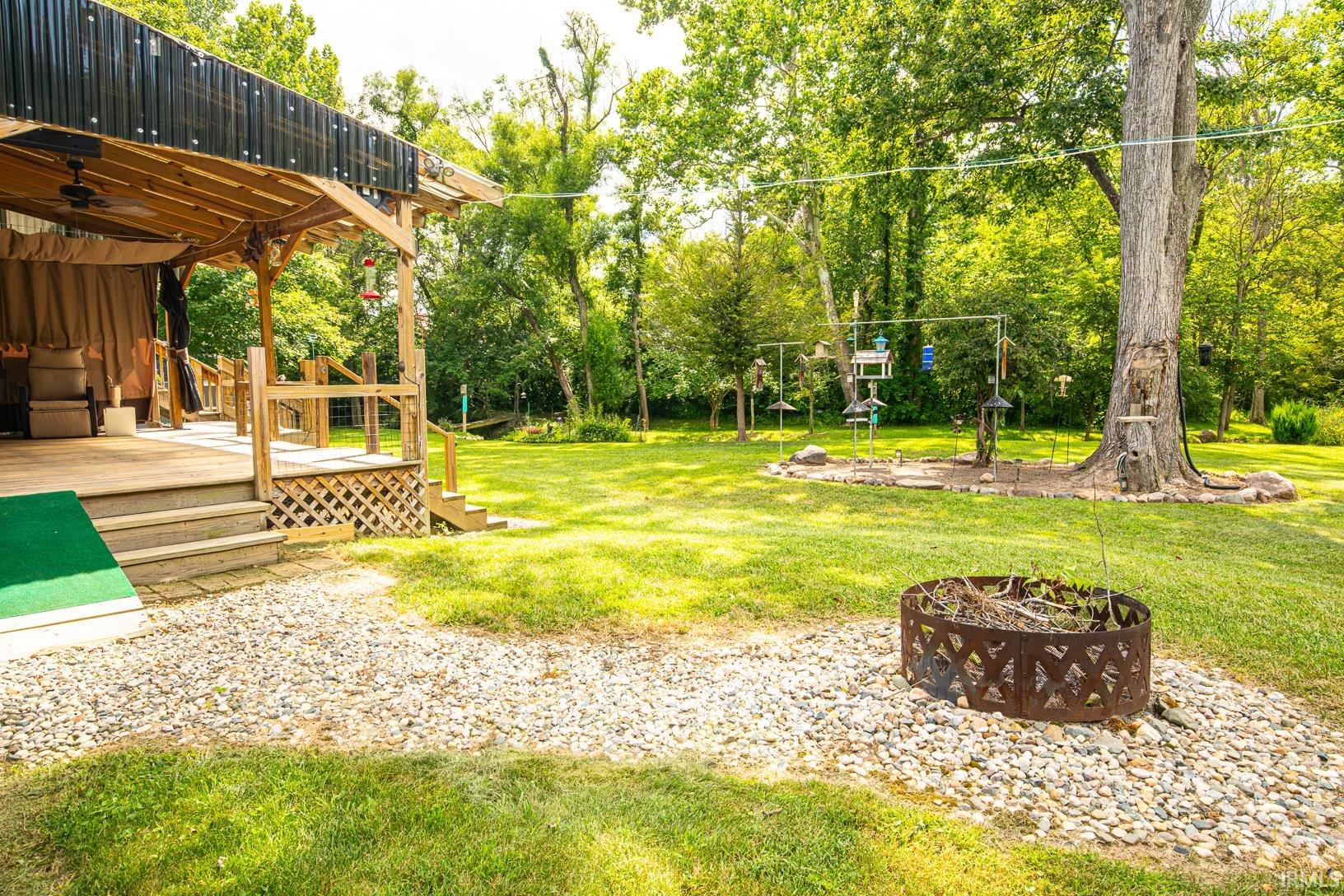 Expansive backyard featuring a covered porch with a corrugated roof, a gravel patio area, a fire pit, mature trees, and a lush lawn