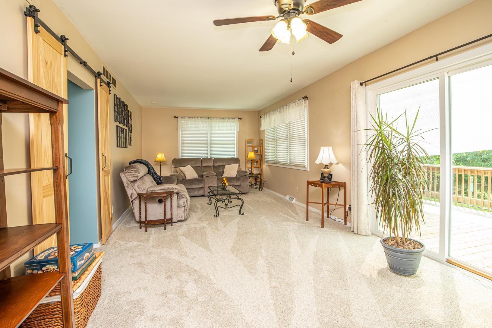 Spacious room featuring light-colored carpeting, a ceiling fan with integrated lighting, and a large sliding glass door opening to an outdoor deck with wooden railing