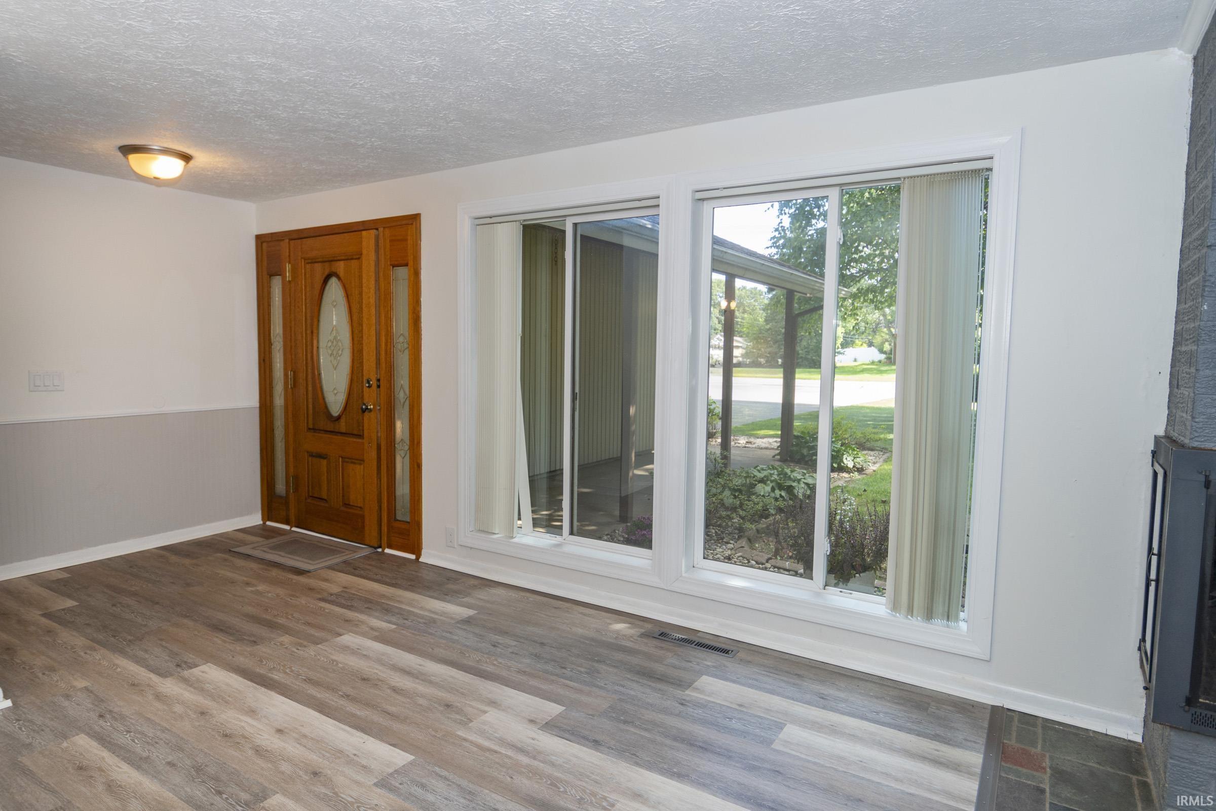 Bright living space featuring wood-finish flooring, an ornate wood entry door with oval glass panel, and expansive sliding glass doors with vertical blinds offering natural light and access to the outdoor grounds