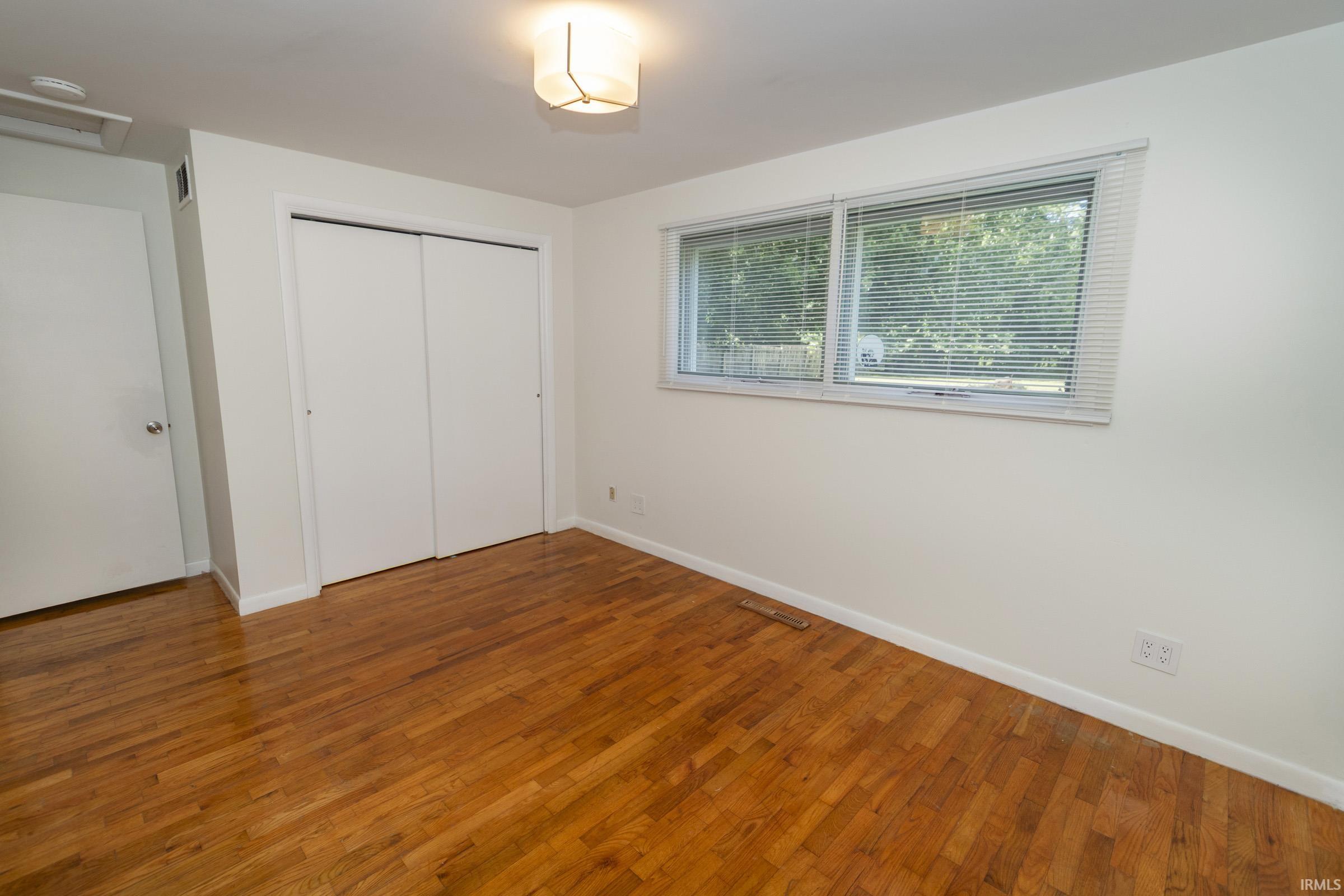 Spacious bedroom showcasing warm wood-finish flooring, white painted closet doors with frame molding, large dual windows with horizontal blinds providing natural light, and a neutral white walls throughout