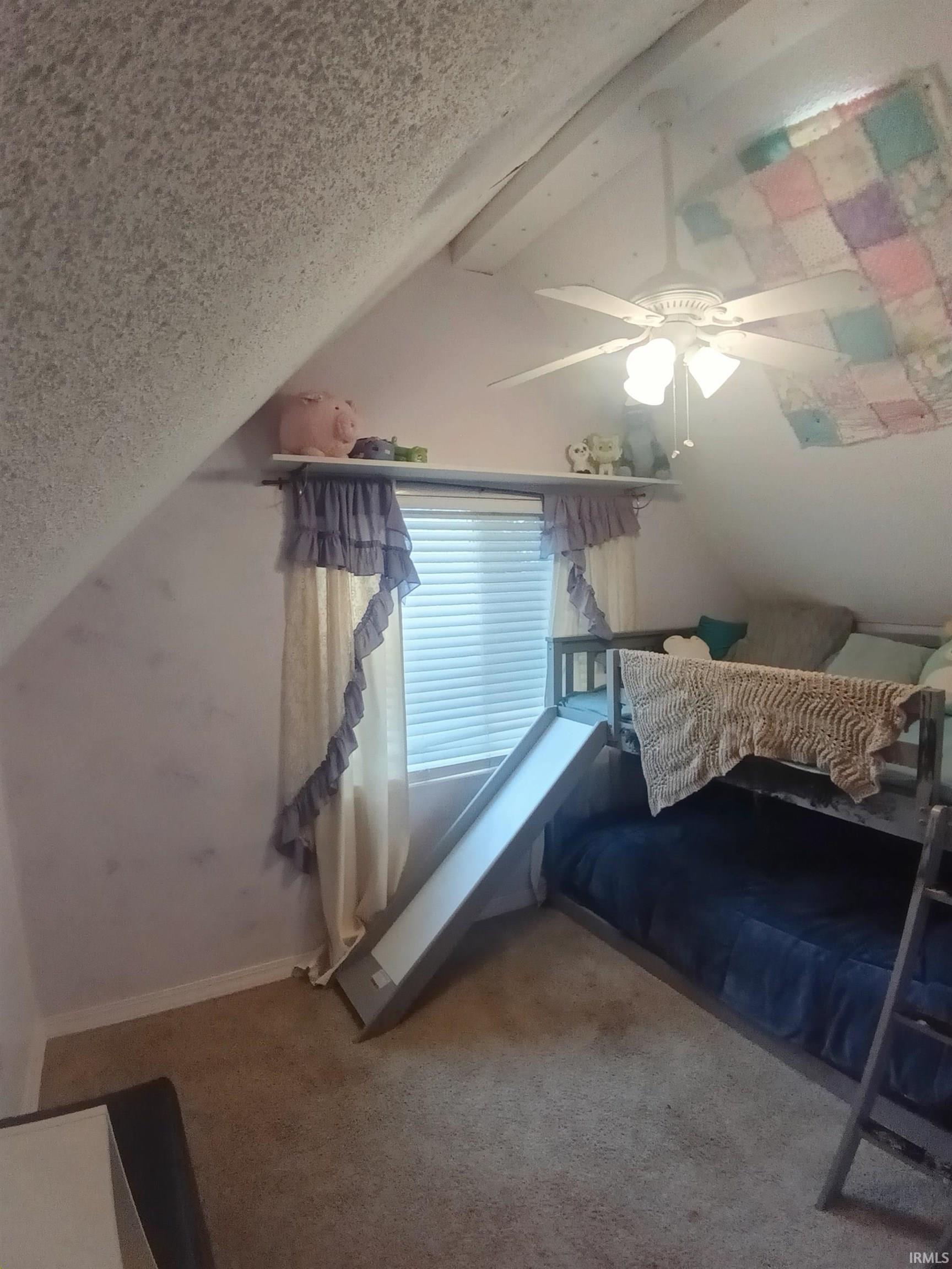 Sloped ceiling with exposed beams, ceiling fan light fixture, wall-mounted shelving, and window with blinds