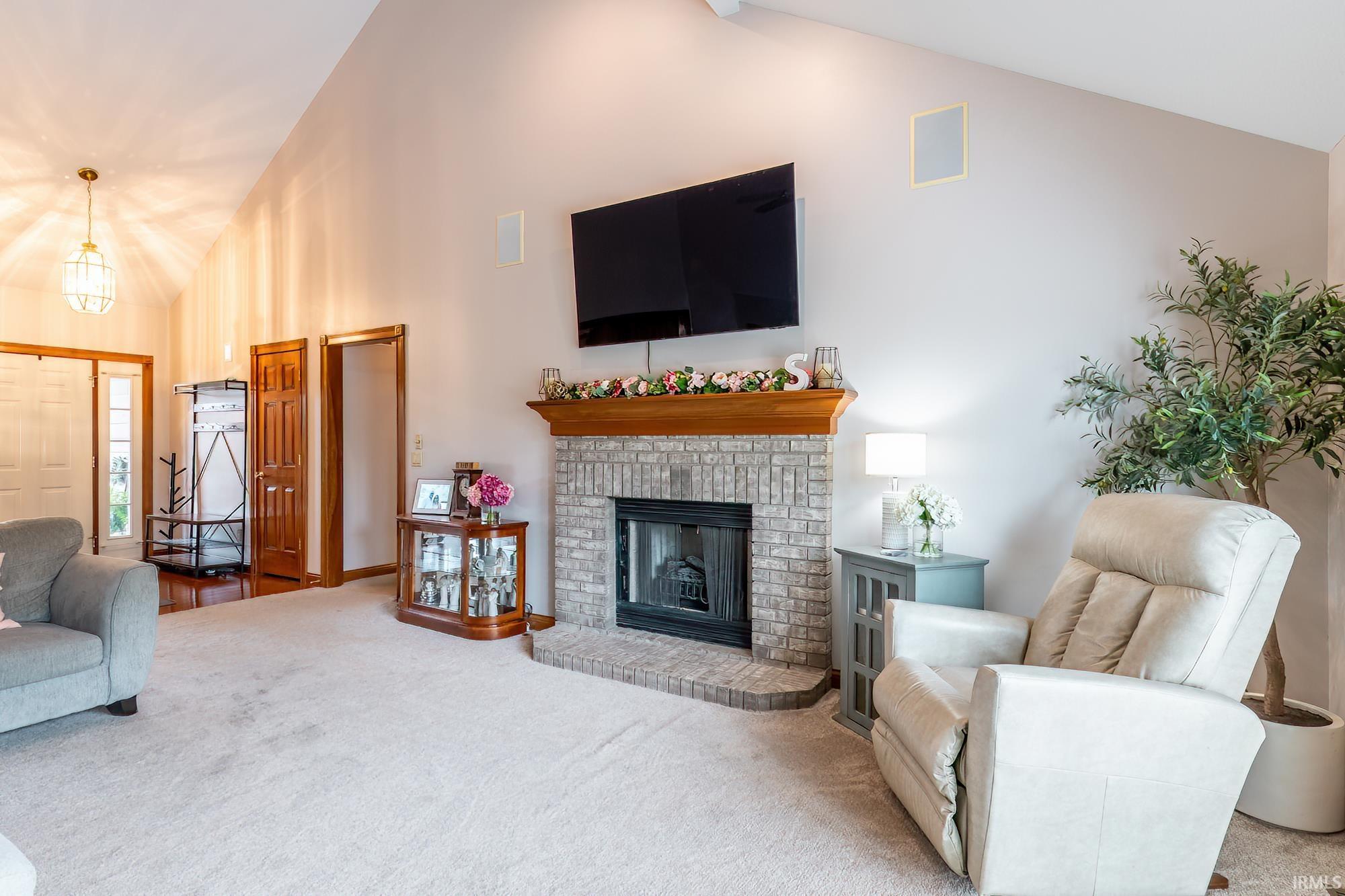 Vaulted ceiling, brick-surround fireplace with a wood mantle, carpeted flooring, and a built-in wall speaker