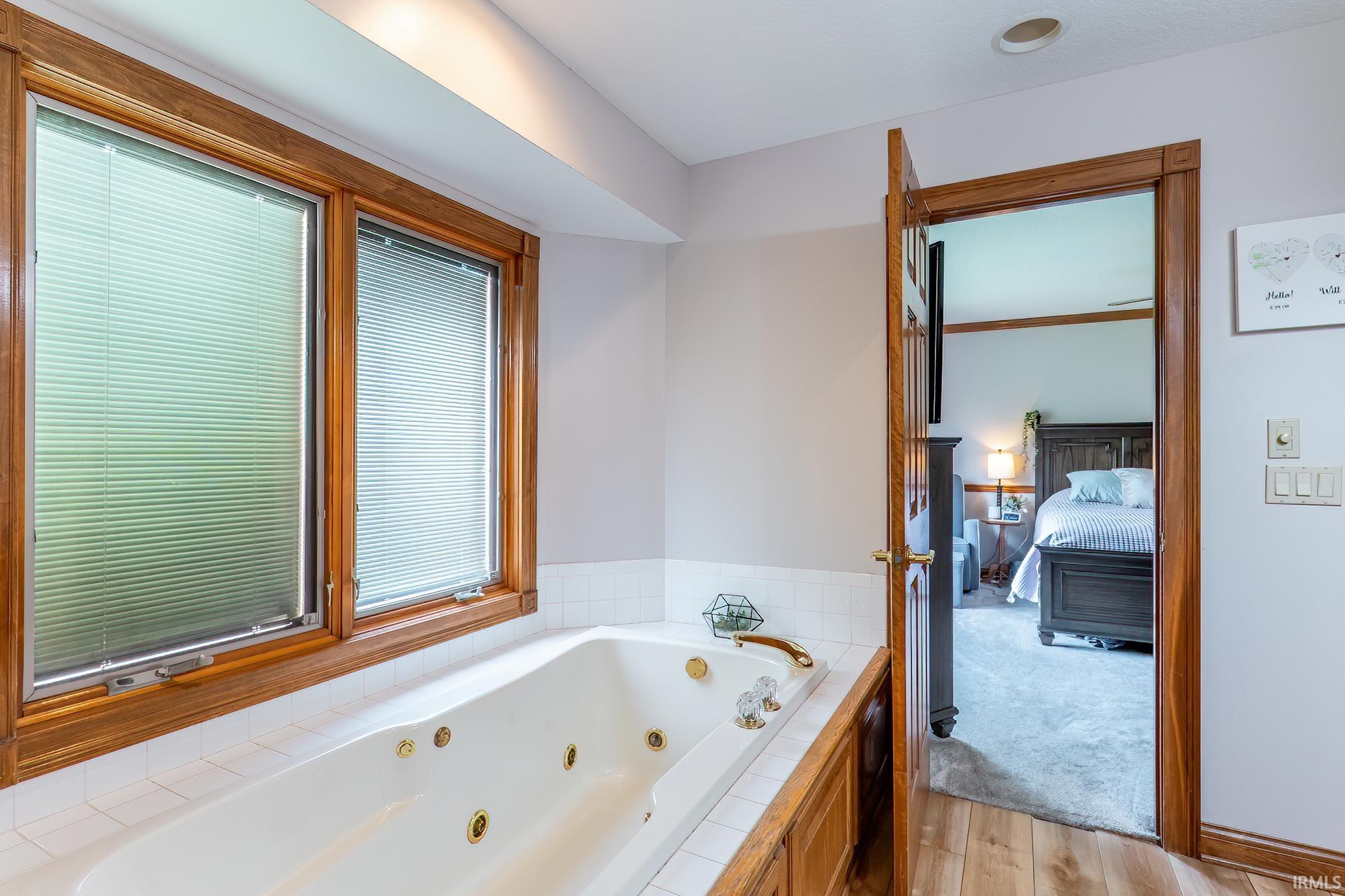 Jetted tub with white tile surround and wood-finish trim, dual windows with blinds, and recessed lighting
