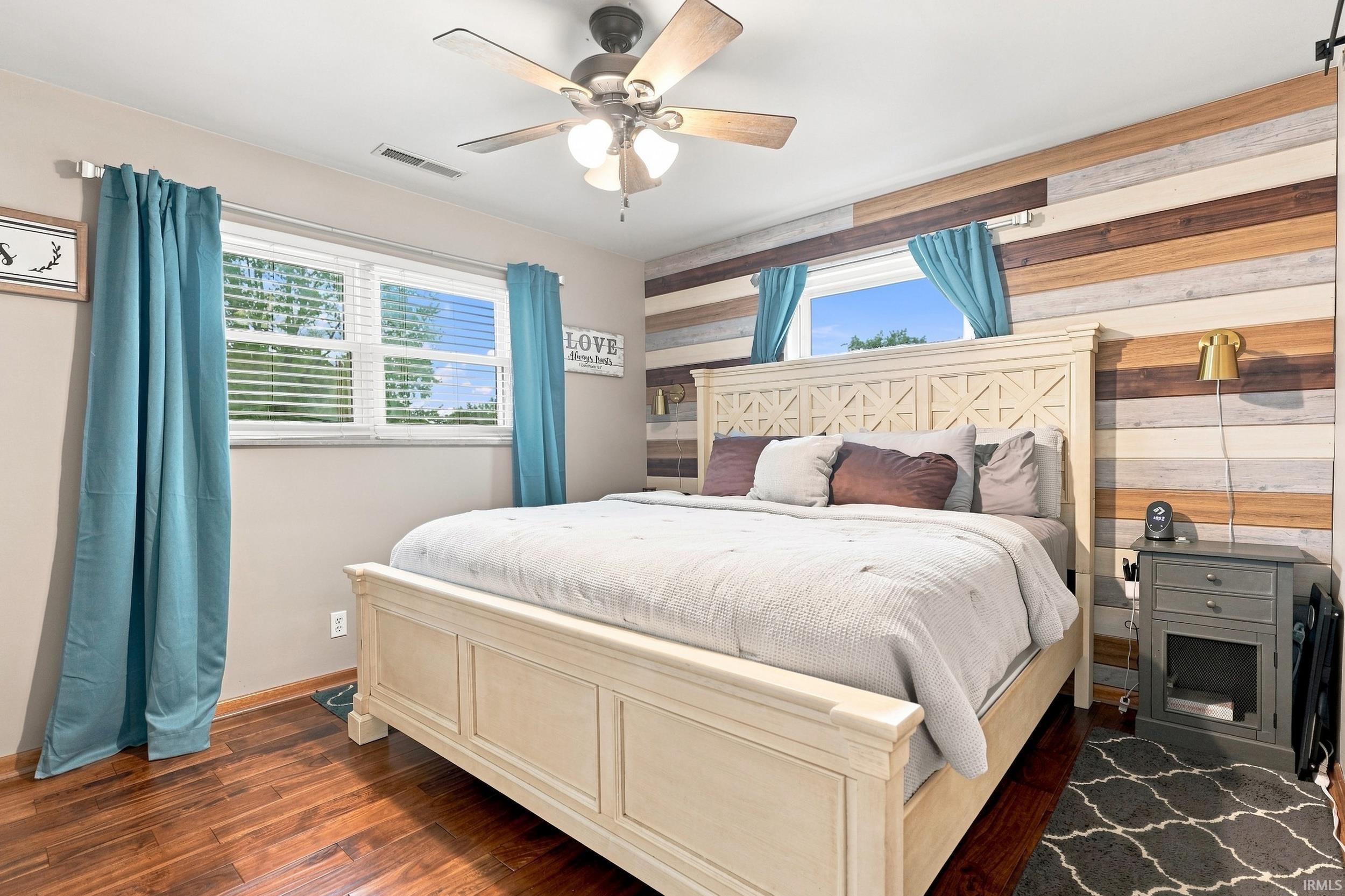 Hardwood flooring, ceiling fan with light fixture, two windows with blinds, and an accent wall with varied wood paneling
