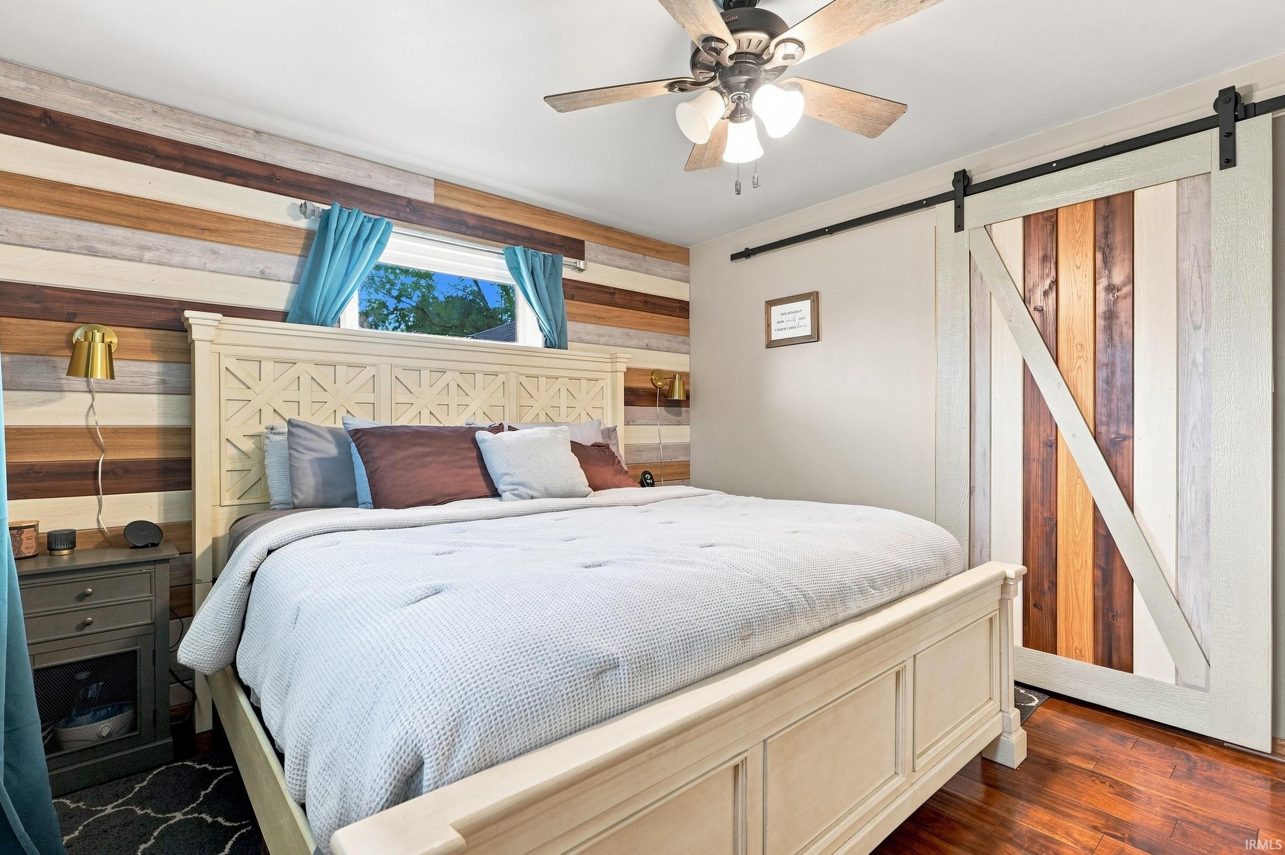 Hardwood flooring, wood-paneled accent wall, ceiling fan, window with natural light, and a barn-style sliding door