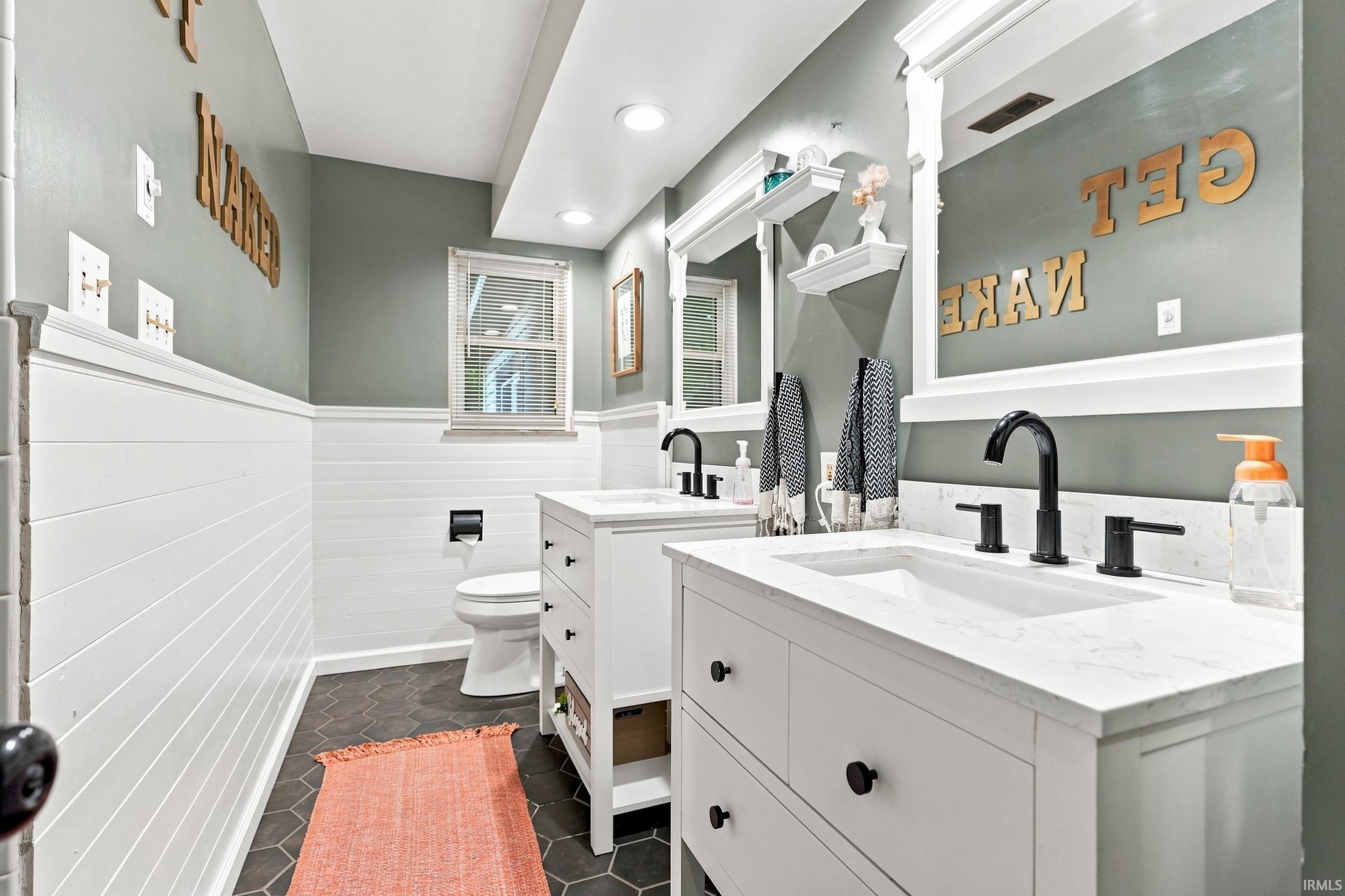 Dual white vanities with integrated sinks, matte black fixtures, shiplap wainscoting, hexagonal floor tiling, and a window providing natural light