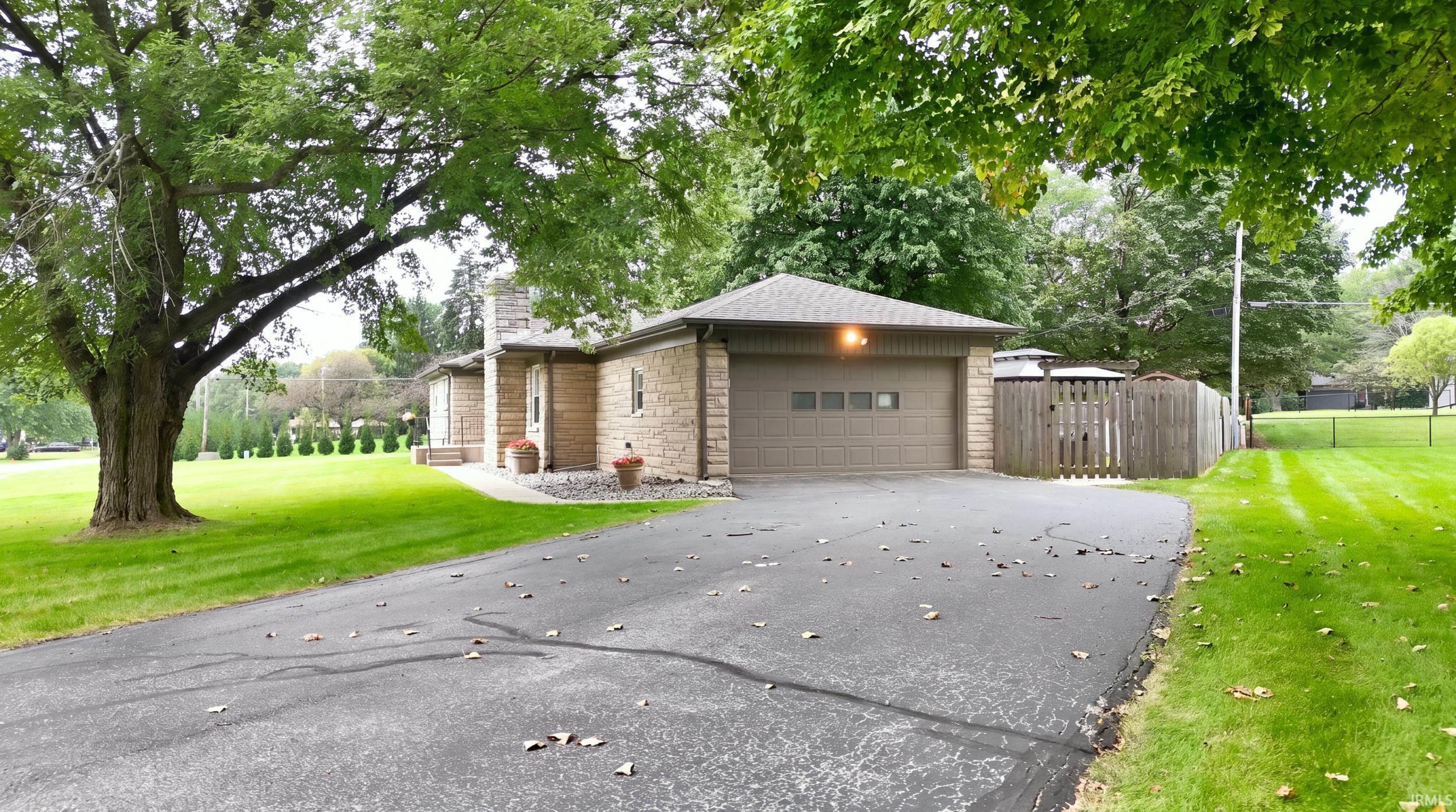 Stone facade residence featuring an attached garage, paved driveway, mature trees, and a wooden privacy fence
