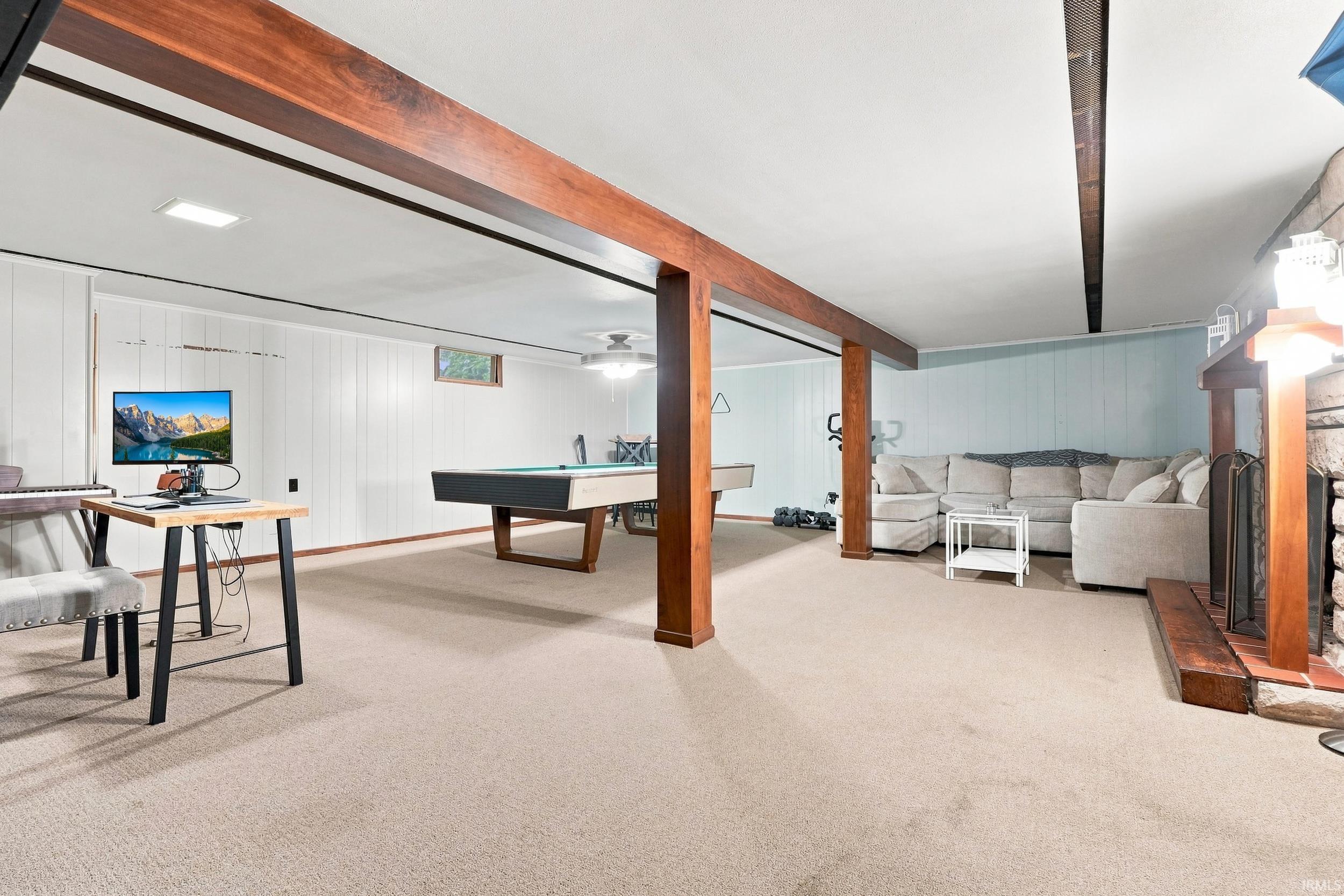 Finished lower level with carpet flooring, wood-finish support beams, stone-finish fireplace, and paneling accents
