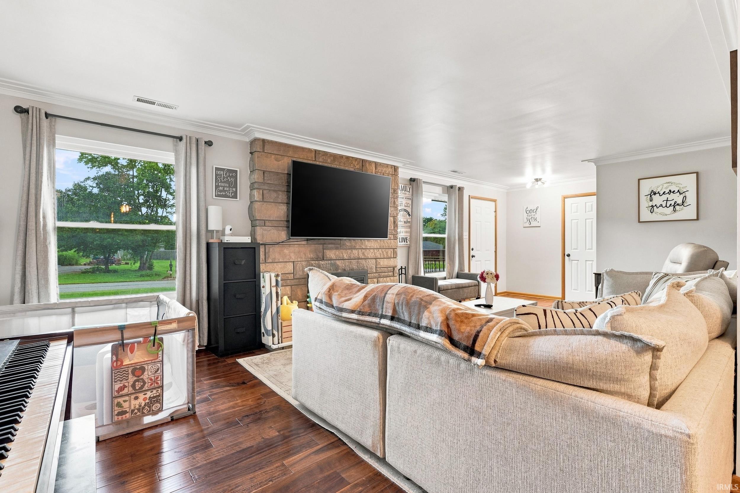 Living area featuring wood-finish flooring, a stone-clad fireplace, white crown molding, and multiple windows