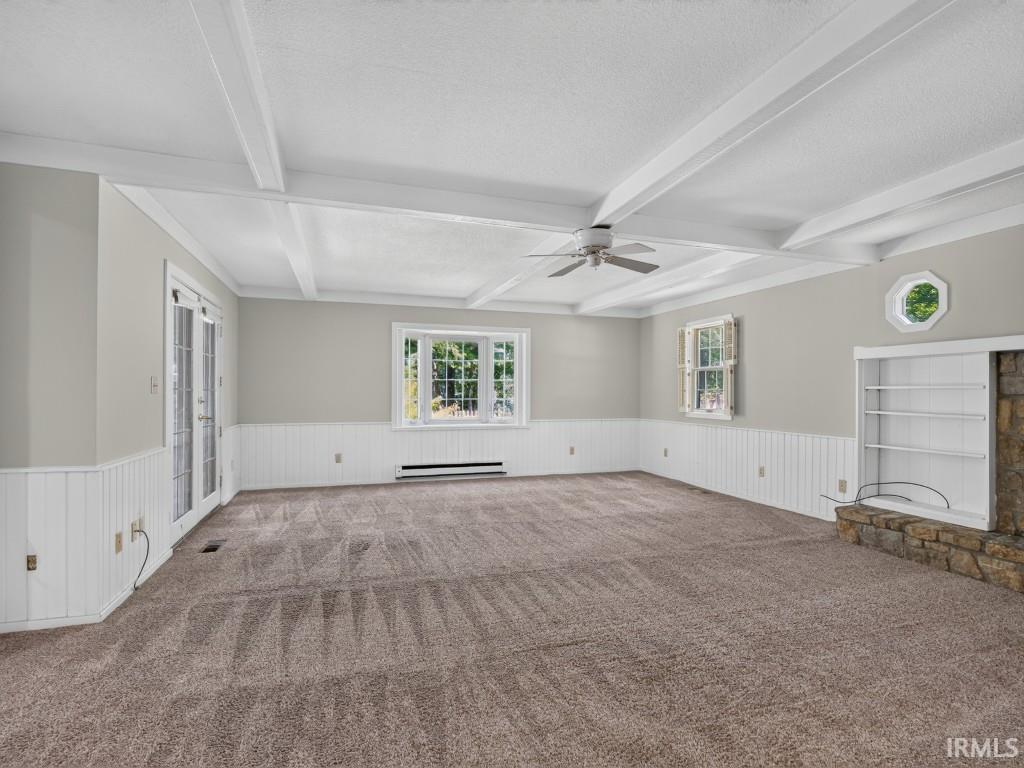 Spacious room with exposed beam ceiling, wall paneling, built-in shelving, stone fireplace surround, and French doors