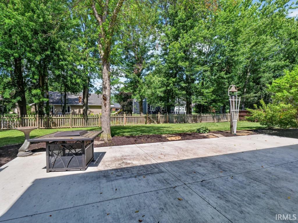 Expansive concrete patio with a fire pit, bordered by a manicured lawn and mature trees, featuring a wood picket fence