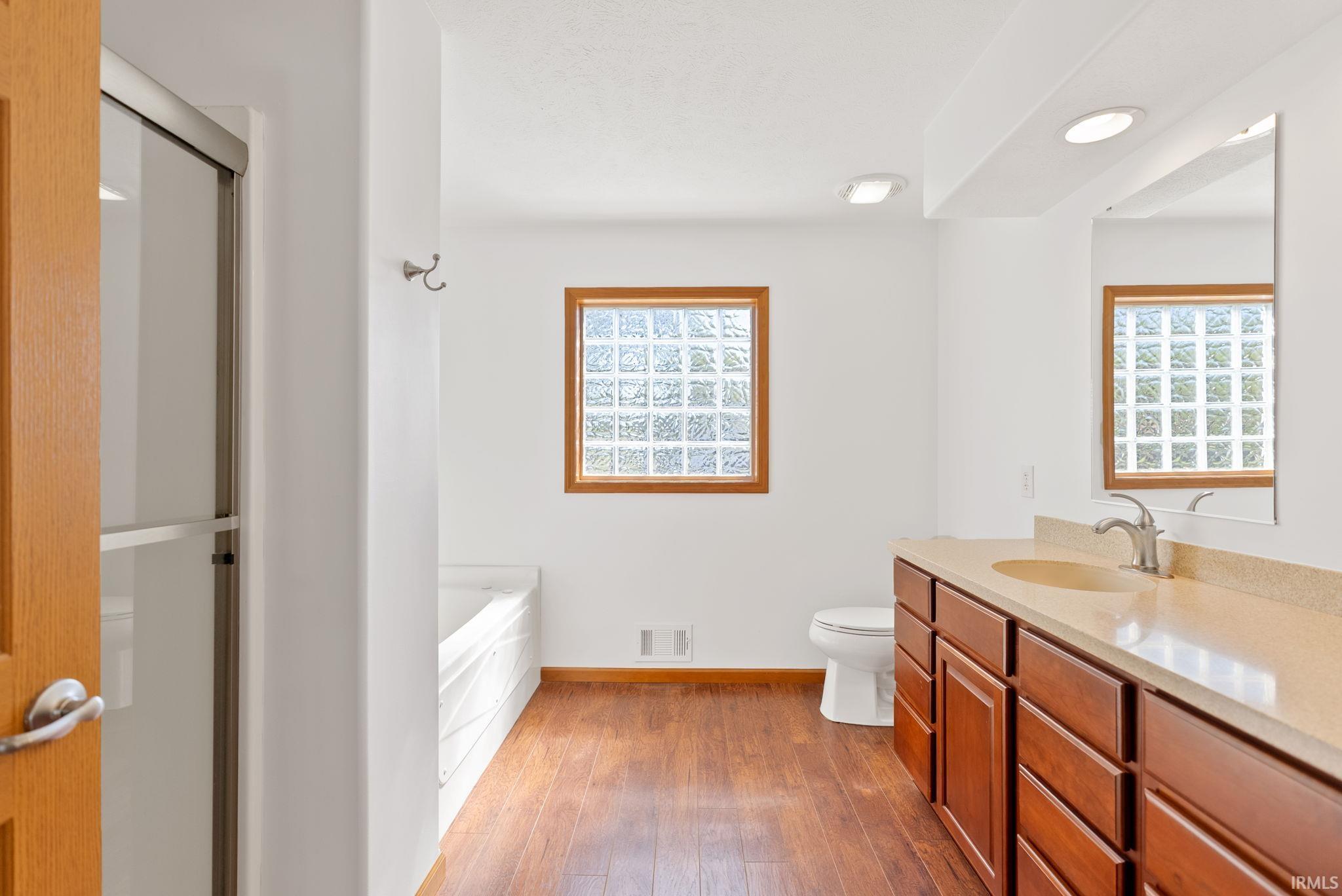 Bathroom featuring wood-finish flooring, a long vanity with dual sinks, warm-toned wood cabinetry, a glass block window, and a built-in tub