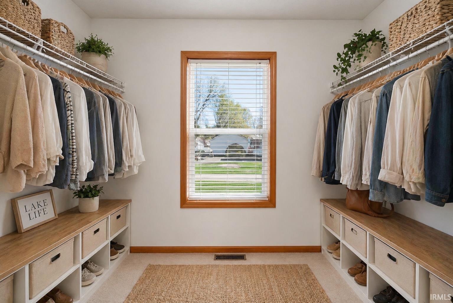 Walk-in closet featuring a centered window with wood trim and horizontal blinds, dual clothing racks with upper shelving, and built-in lower storage units with wood-finish tops
