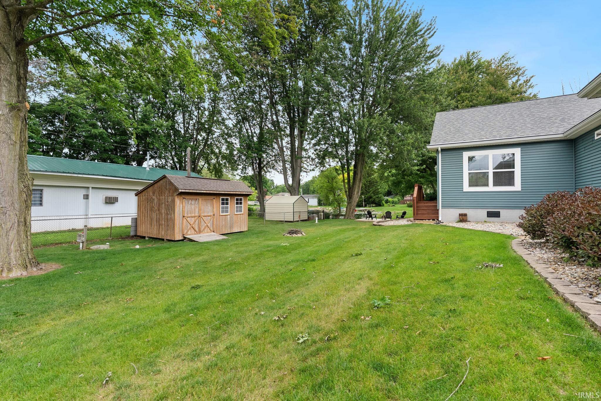 Expansive green lawn with a newly constructed wood-sided shed, mature trees, a fire pit, and a portion of the residence featuring blue siding and white window trim
