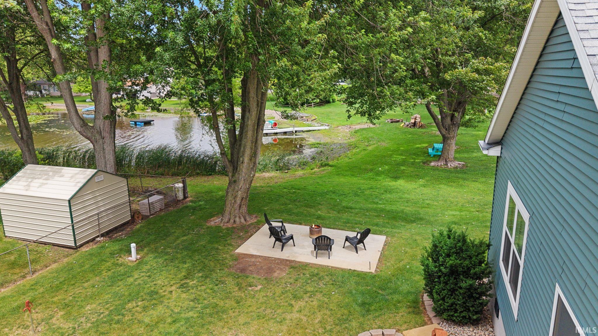Expansive green space featuring mature trees, a concrete patio, a storage shed, and a body of water with a dock