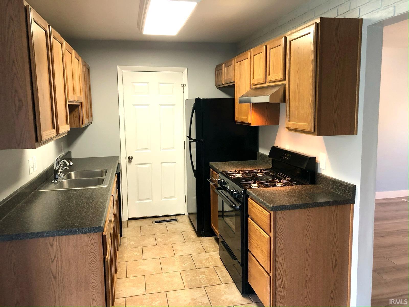 Kitchen with wood-finish cabinetry, dark countertops, tile flooring, and a stainless steel double basin sink
