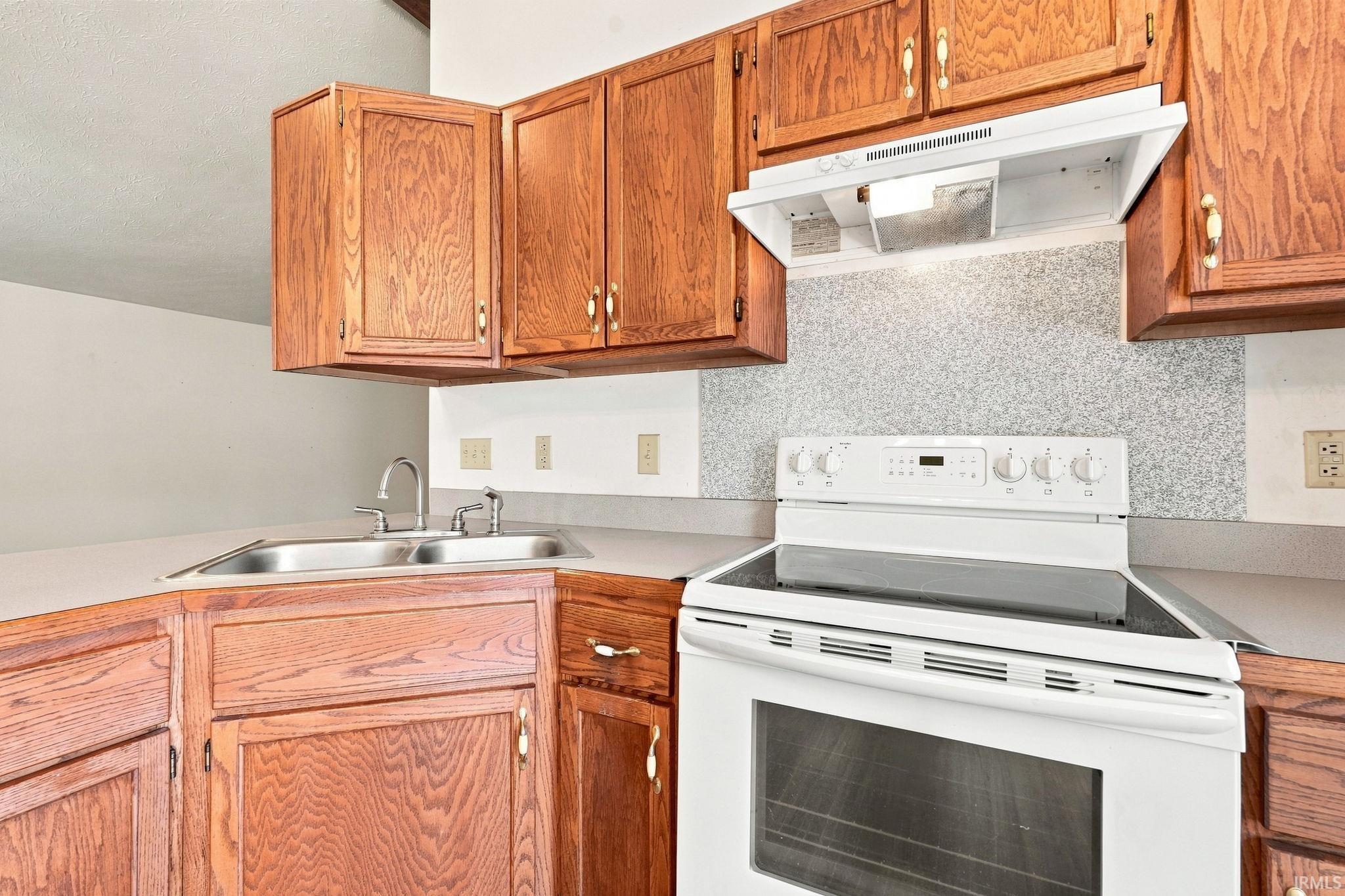 Wood-finish cabinetry, corner stainless steel double sink, gray countertop, white electric range, and range hood
