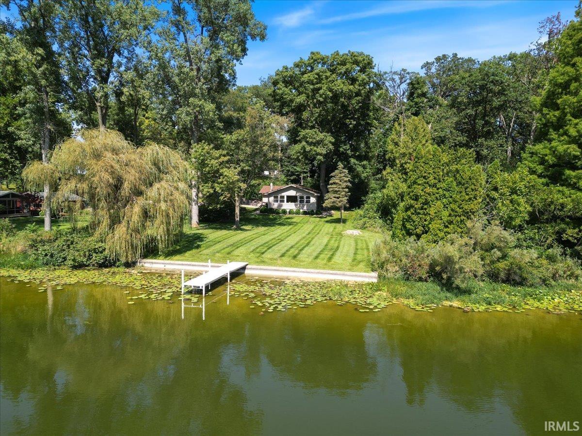 Waterfront property featuring a private dock, expansive green lawn, mature trees, and a visible single-story residence