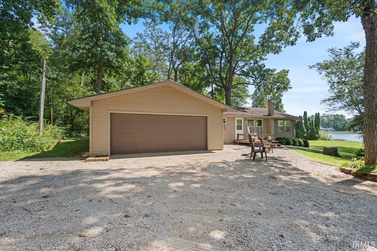 Detached garage with a roll-up door, gravel driveway, and a single-story residence with a pitched roof, rear patio, and a grass lawn extending to the water