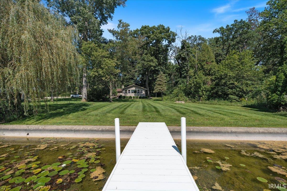 Private dock extending over a waterway, verdant lawn, mature trees, and a single-story residence