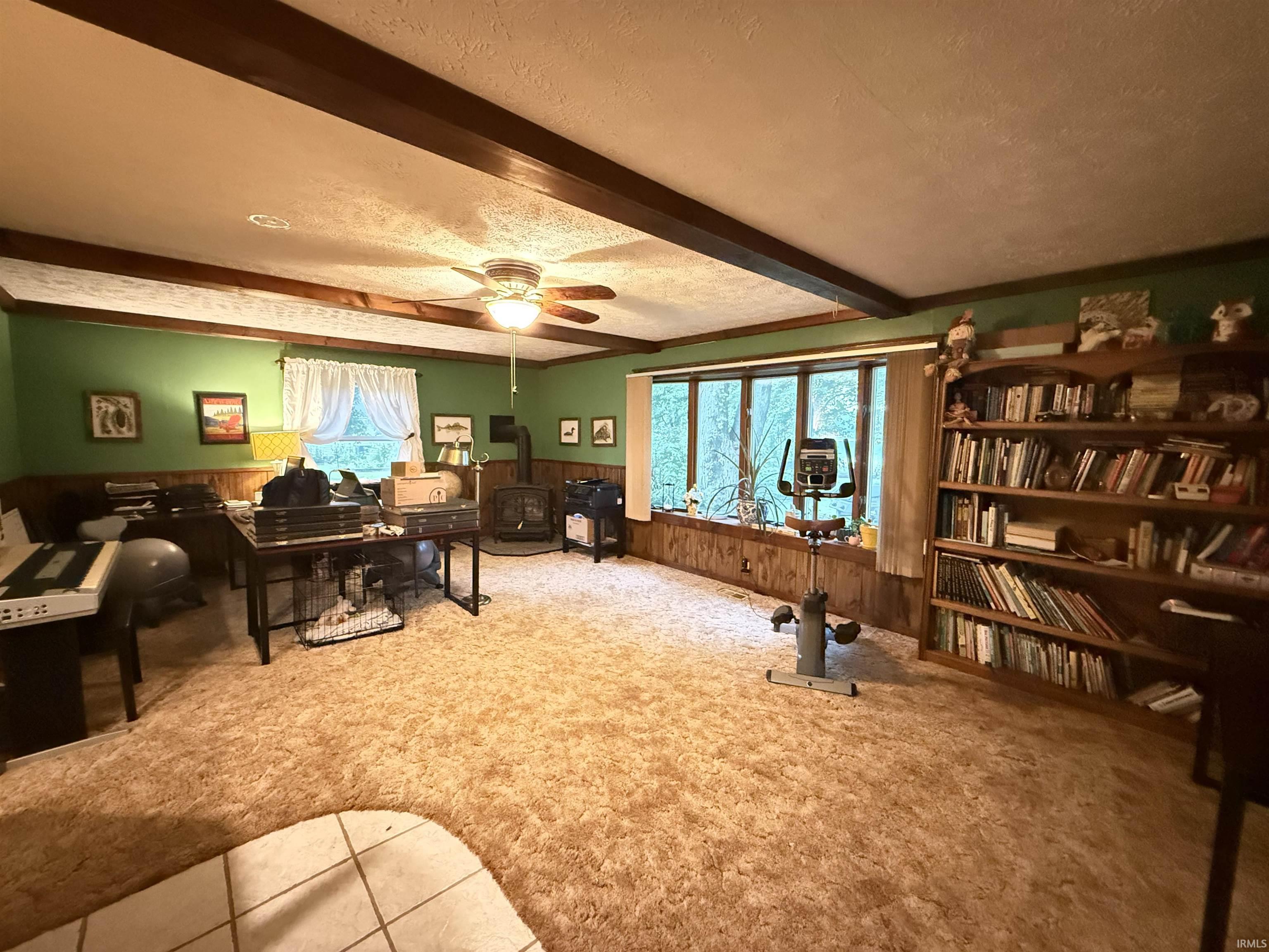 Spacious room featuring wood-finish ceiling beams, a ceiling fan with integrated lighting, wood-paneled wainscoting, and a built-in wood bookshelf